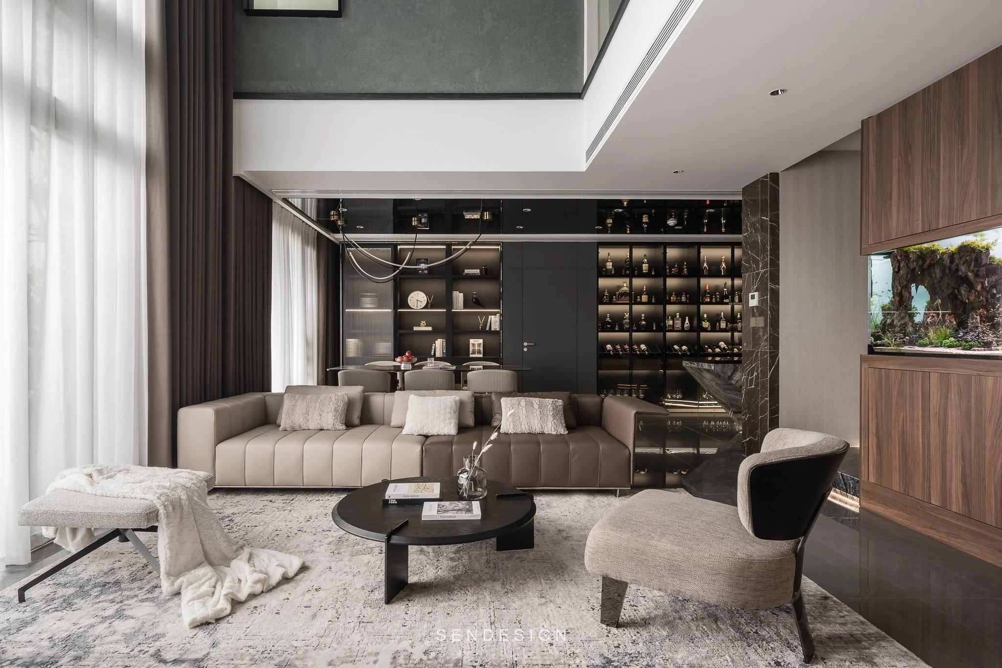 Modern living room with beige sofa, black coffee table, and framed photos. Large window with white curtains. Bookshelf with decorative items and bottles. Rug on the floor.