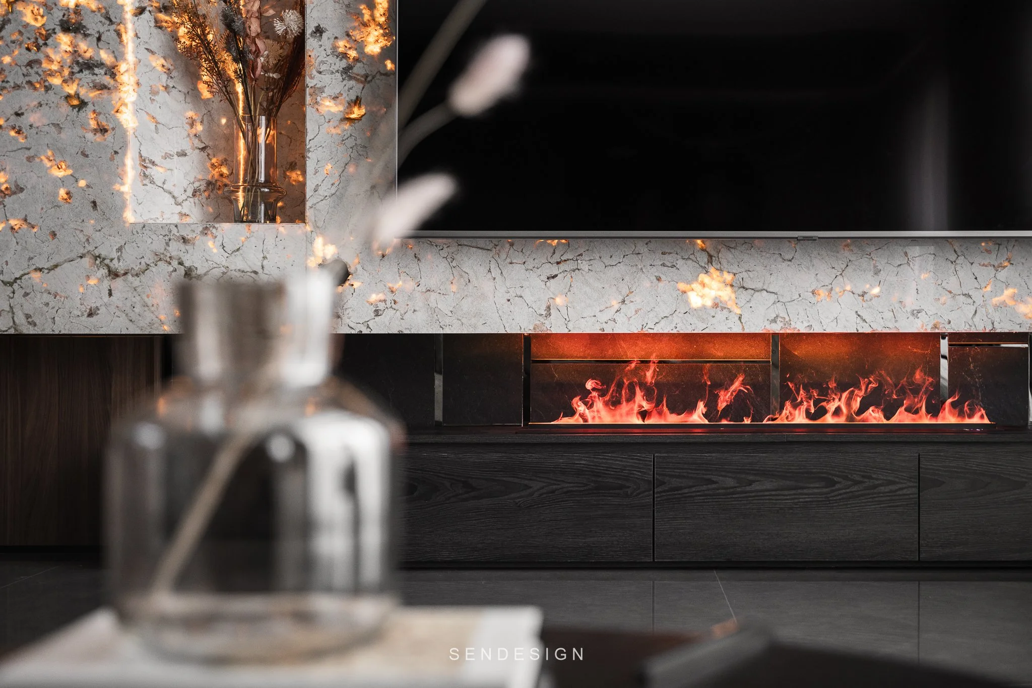 A modern fireplace with a fiery orange flames design under a black screen, set against a marble wall with gray veins, and a blurred silver object in the foreground.
