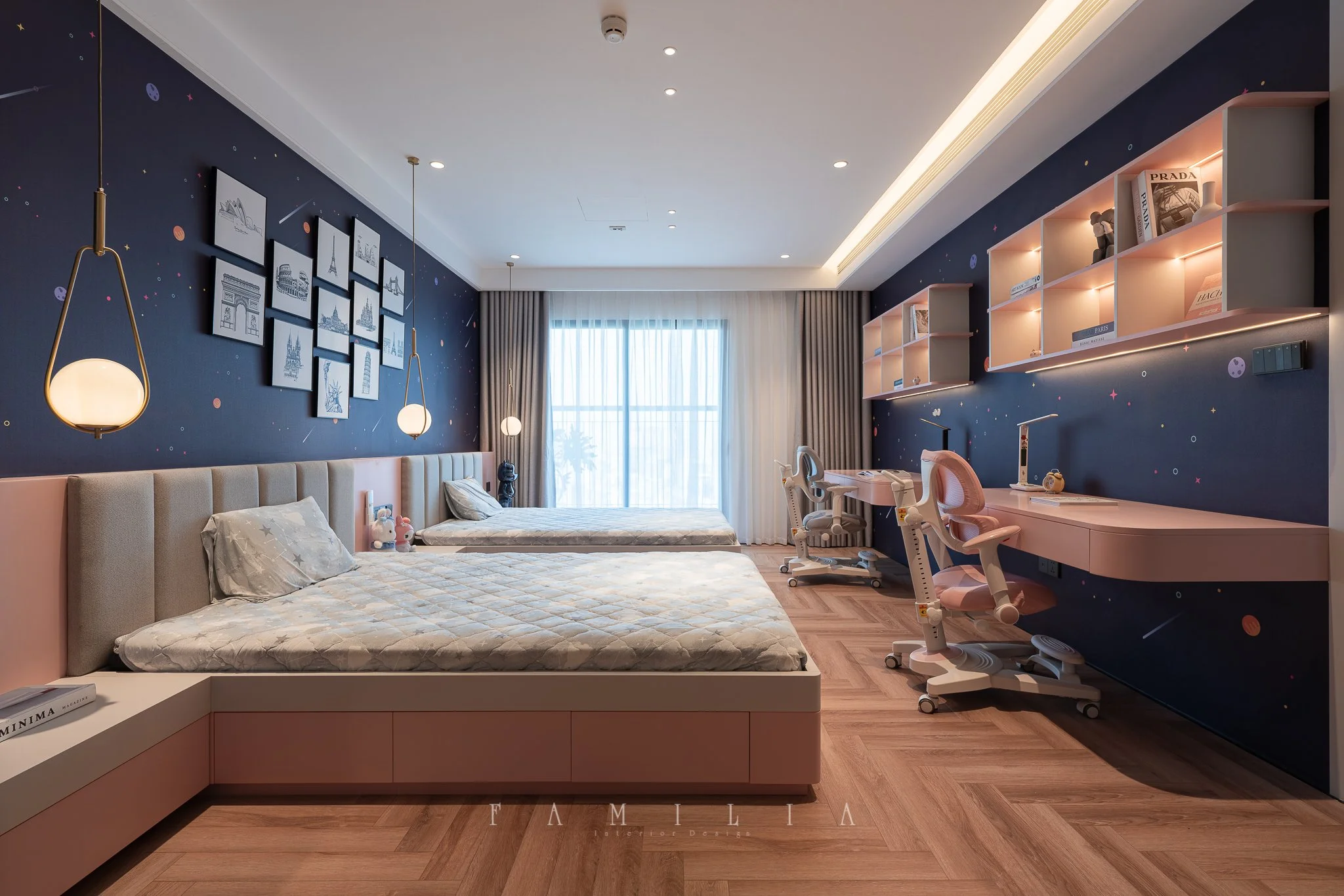 A modern bedroom with two beds, a blue celestial-themed wall with framed artwork, a beige headboard, and pink furniture. There are pendant lights hanging from the ceiling, a large window with curtains, and a desk with chairs and study lamps. Shelves 