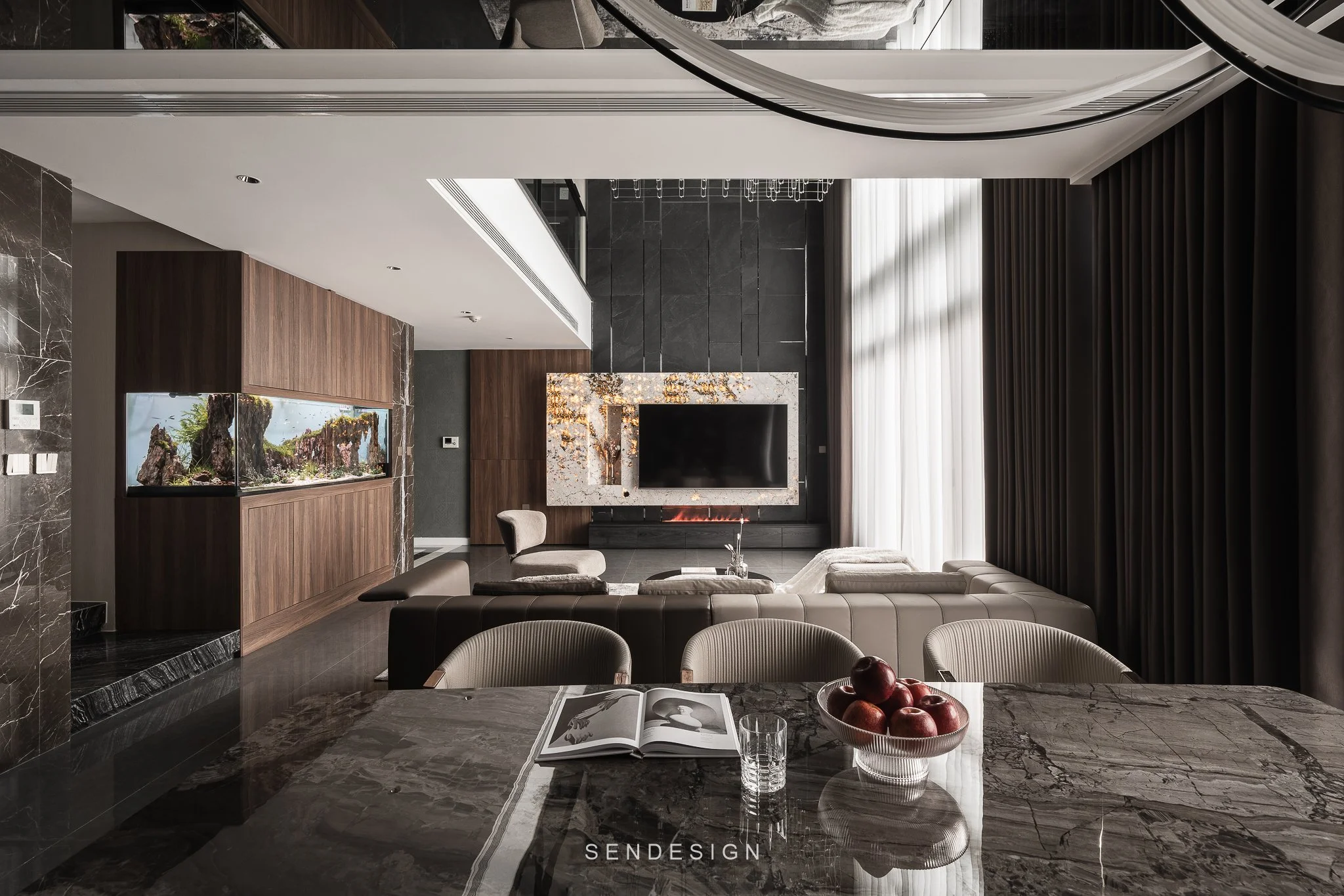 Modern living room with black marble flooring, a fireplace with a marble mantel, a flat-screen TV, a large fish tank, and a white sofa. Bright sunlight streams through large windows with white curtains, and a bowl of apples is on the marble dining ta