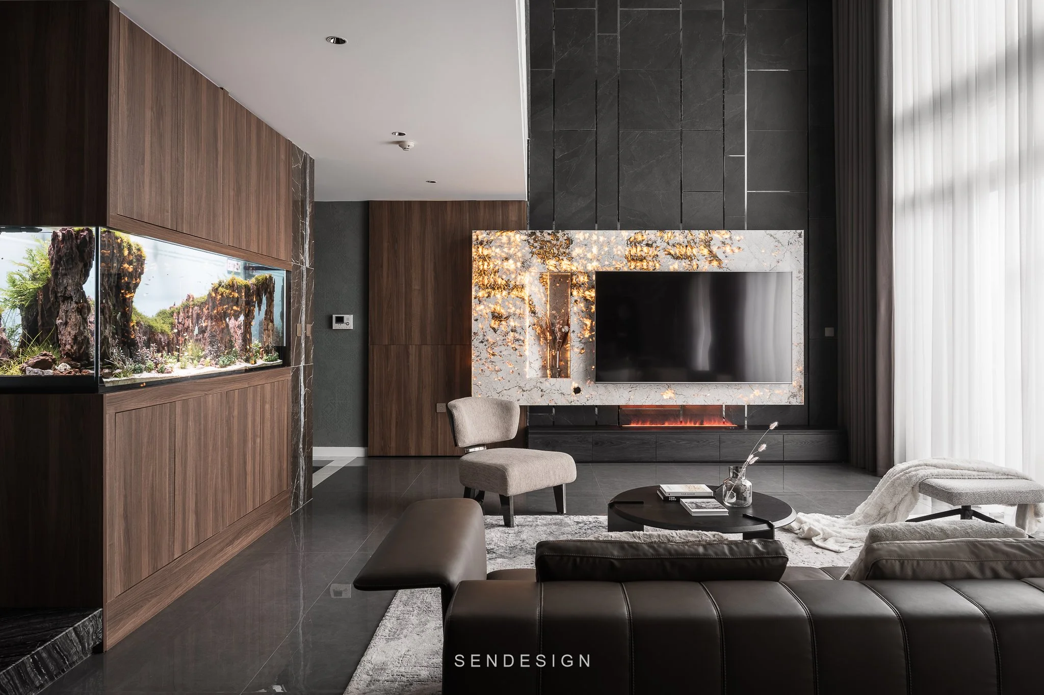 Modern living room with black tiled walls, a marble fireplace with lighting, a large flat-screen TV, and seating including a beige armchair and black leather furniture. There's a large window with sheer curtains on the right.