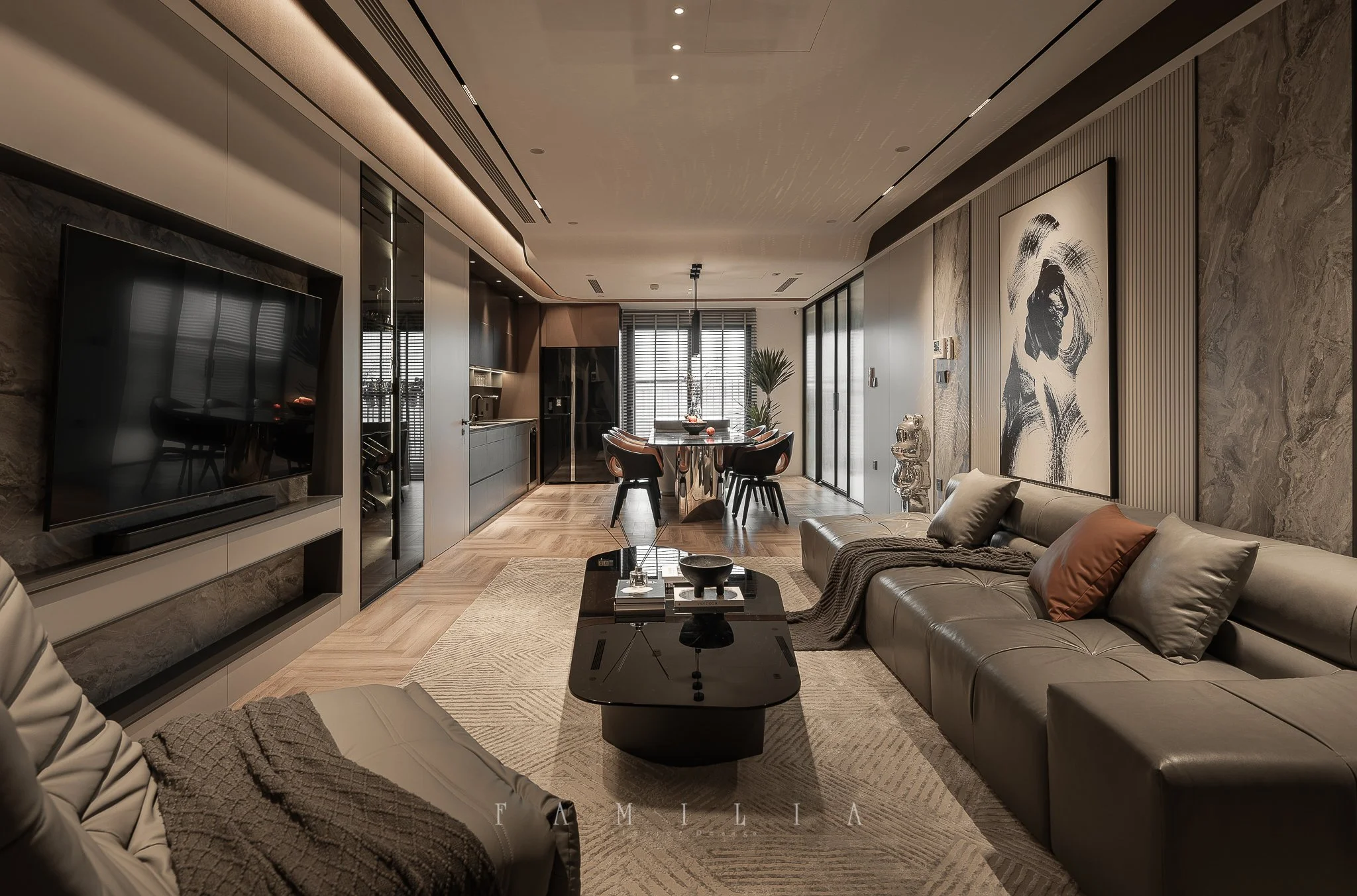 Modern living room with a gray leather sofa, black coffee table, wall art, and a dining area with chairs and table in the background. There is a large flat-screen TV on the wall and natural light coming from windows with blinds.