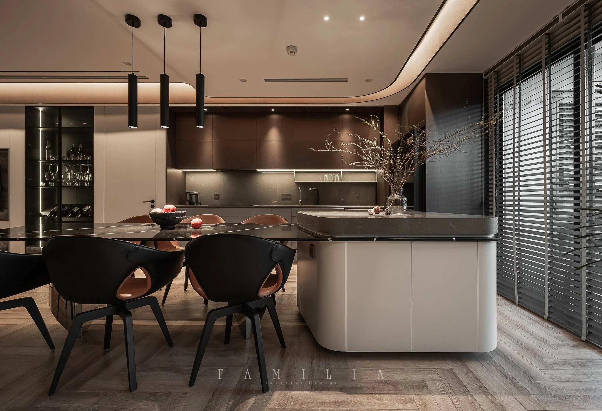 Modern kitchen with dark wooden cabinets, a large island, black pendant lights, a glass cabinet with wine and glasses, a vase with branches, and horizontal window blinds.