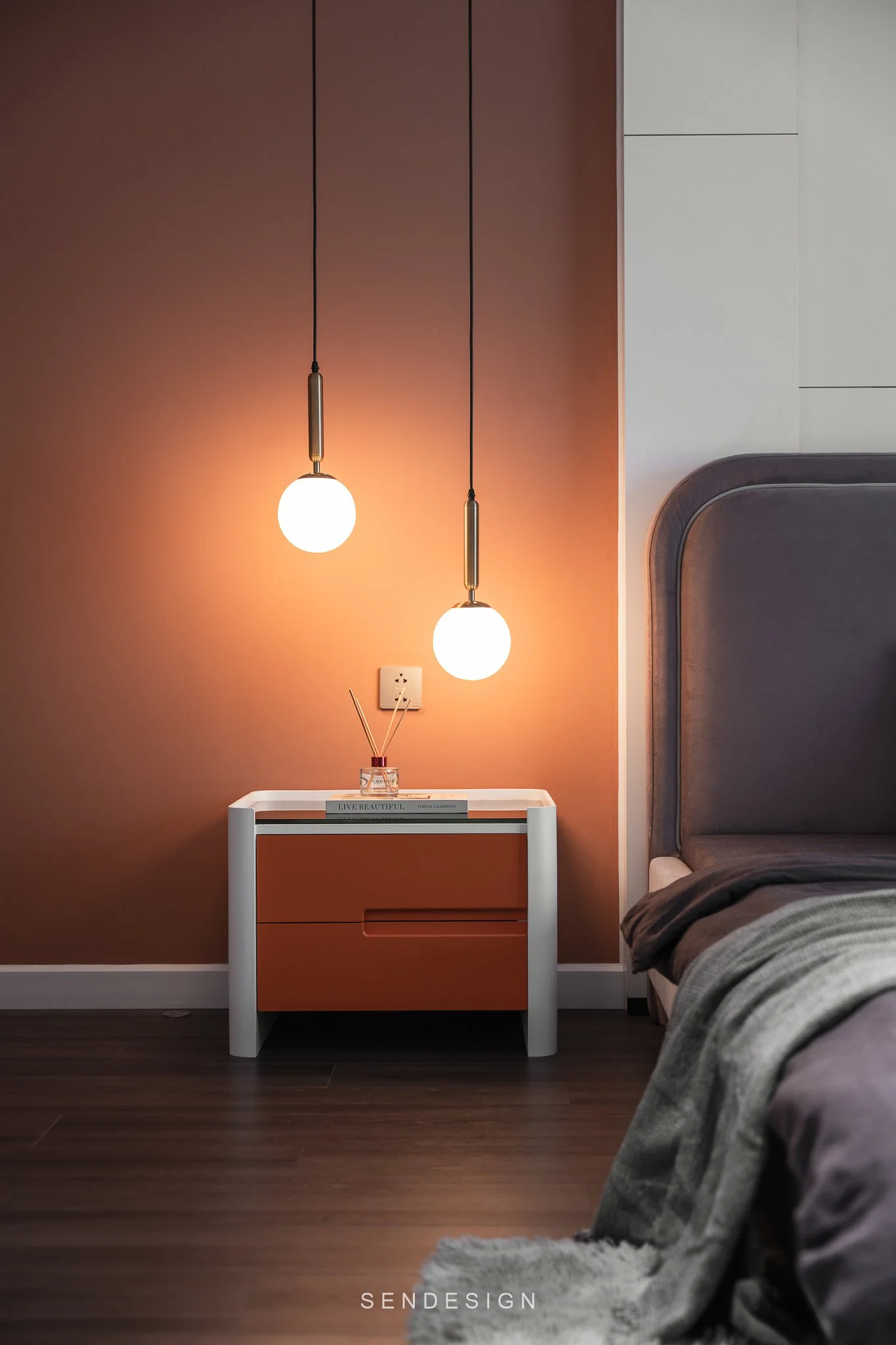 Modern bedroom with a brown accent wall, two hanging globe pendant lights, and a white nightstand with a small bouquet of reed diffusers and a book titled 'Live Beautiful.' Part of a bed with gray and brown bedding and a gray upholstered headboard vi