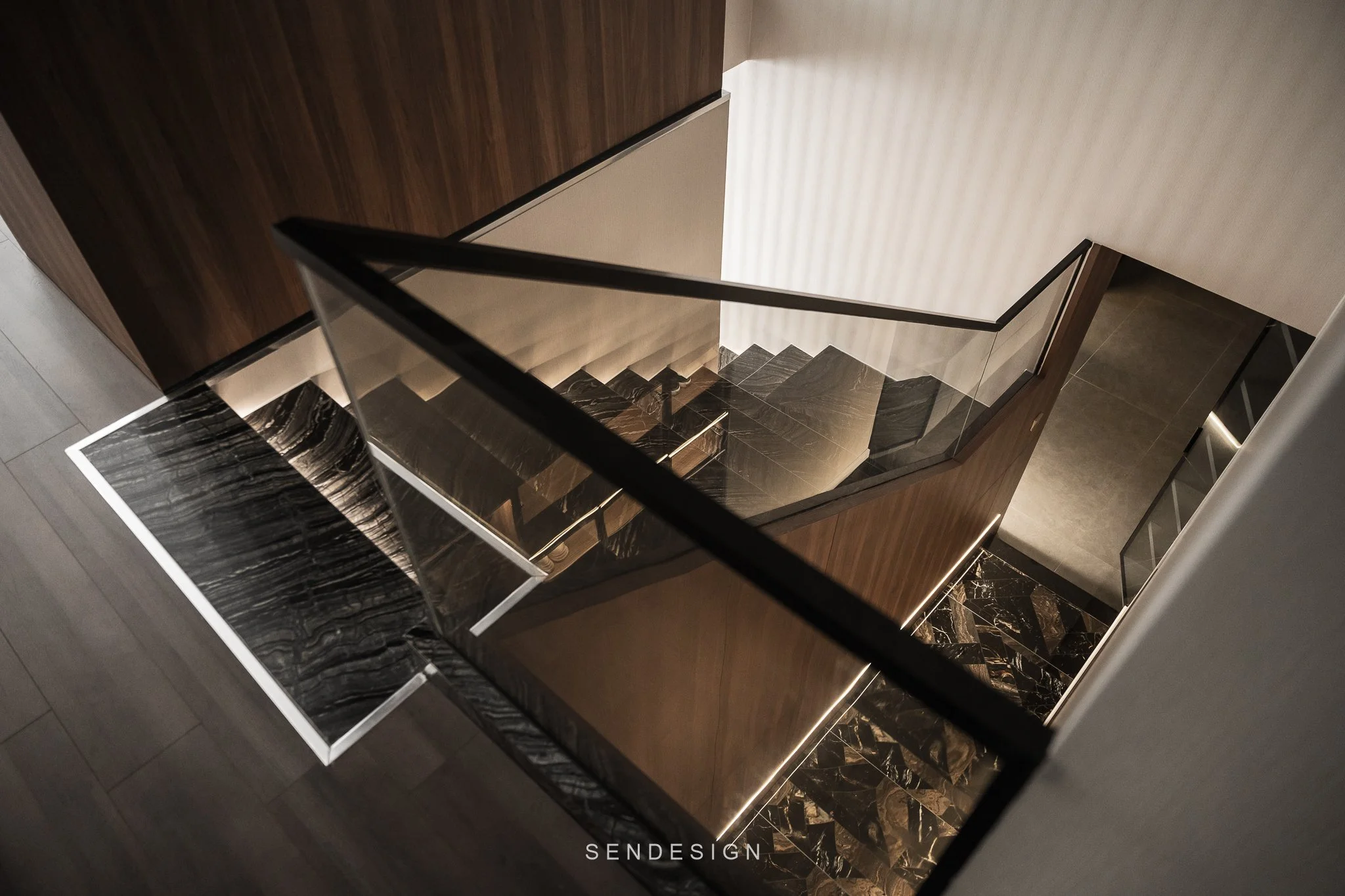 View from the top of a staircase showing dark wooden and black marble steps, glass railing, and a patterned beige wall.