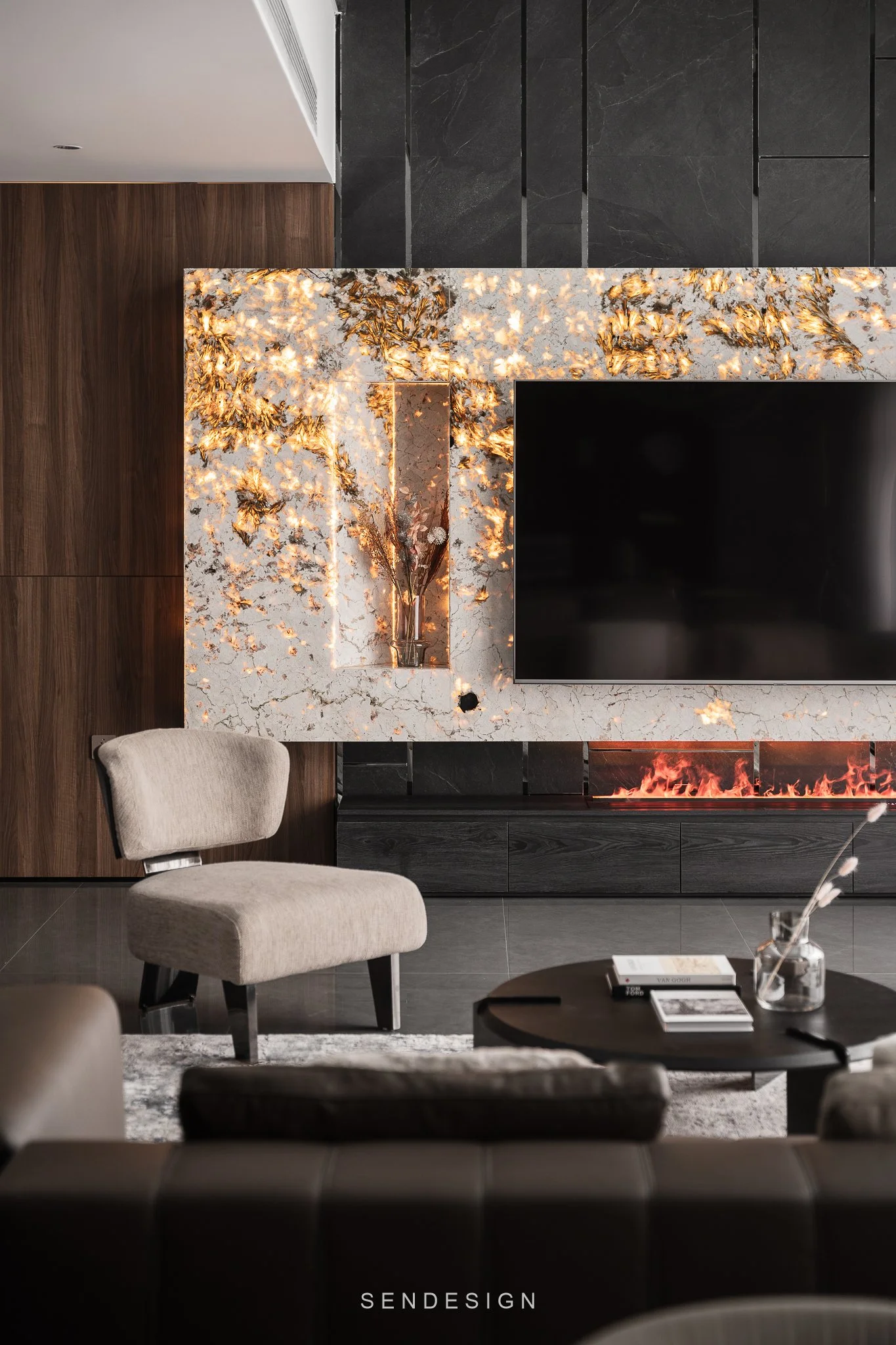 Modern living room featuring a marble fireplace with gold accents, a black flat-screen TV mounted above, dark wood paneling, black wall tiles, a beige upholstered armchair, a black round coffee table with books and a glass vase with flowers, and a gr