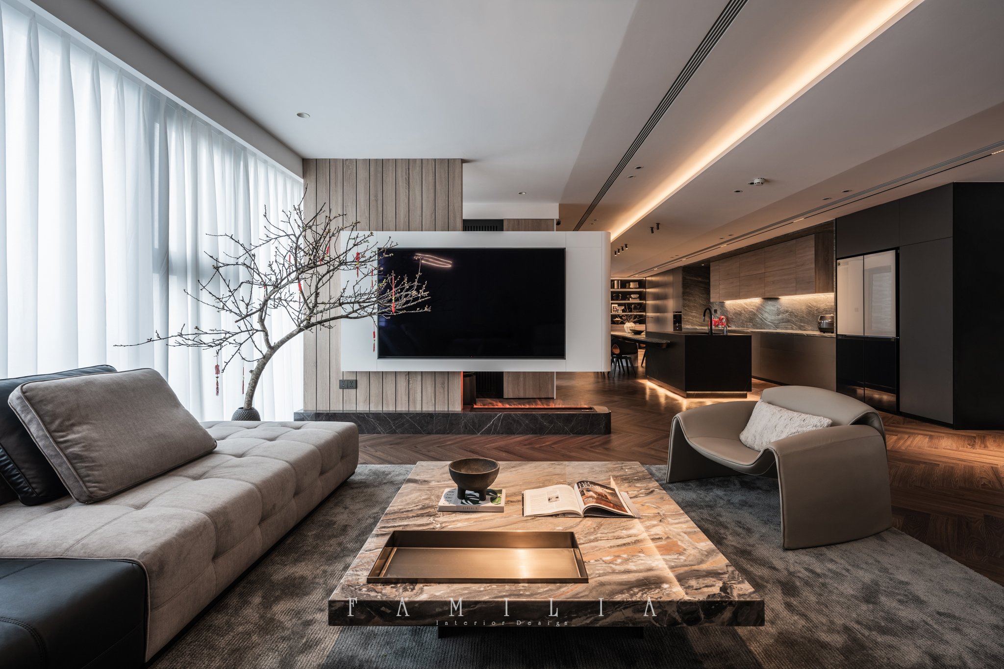 Modern living room with beige sofa, armchair, wooden coffee table with open book, decorative tray, and tray with the word 'FAMILIA' on it. A tree sculpture with red accents is next to a large flat-screen TV mounted on a white wall with wood paneling.