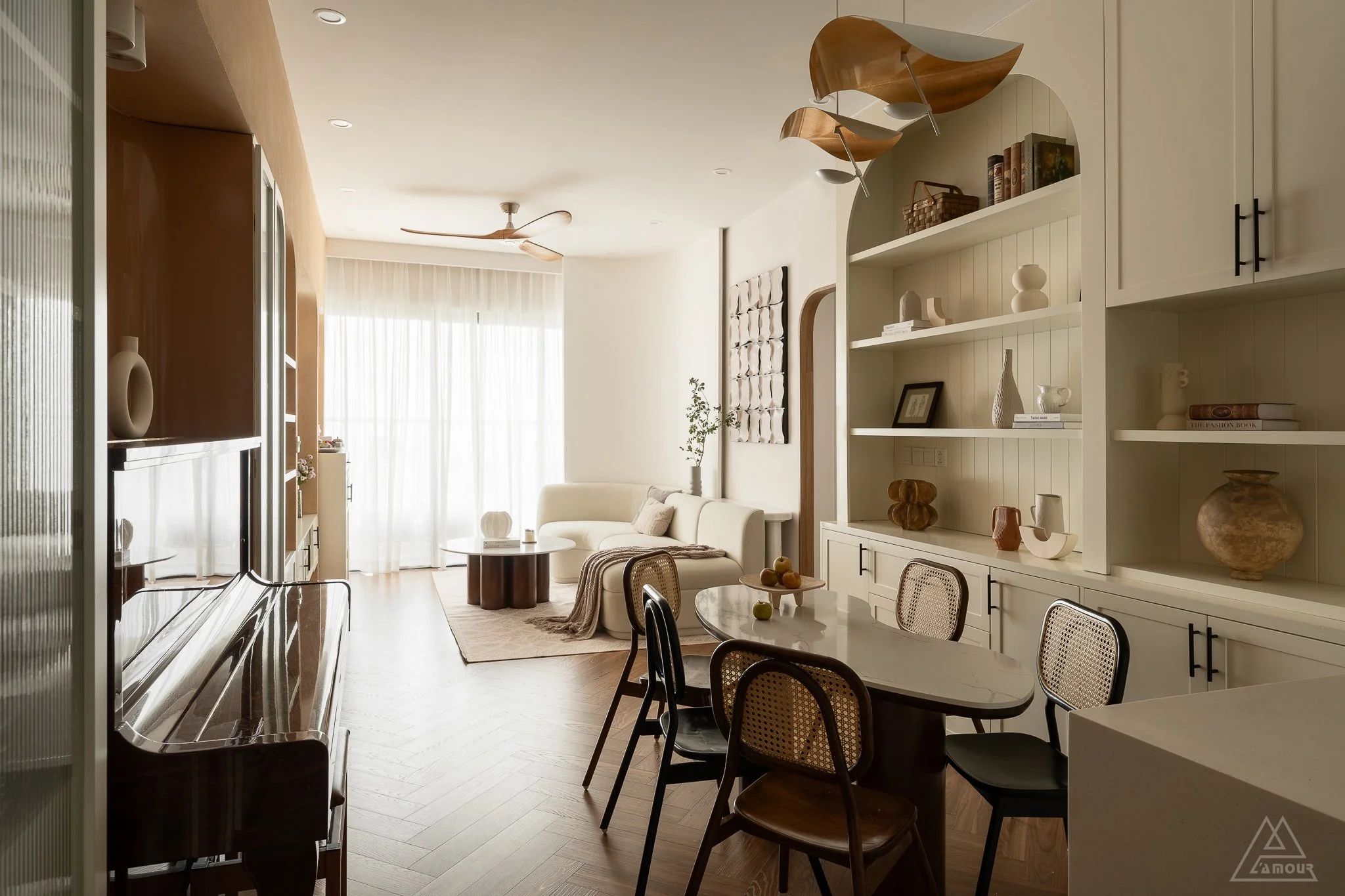Living room with a white sofa, wooden coffee table, and a textured wall art. Large window with curtains, ceiling fan, and built-in white shelves with books and decorative items. Dining table with chairs, and a wooden piano.