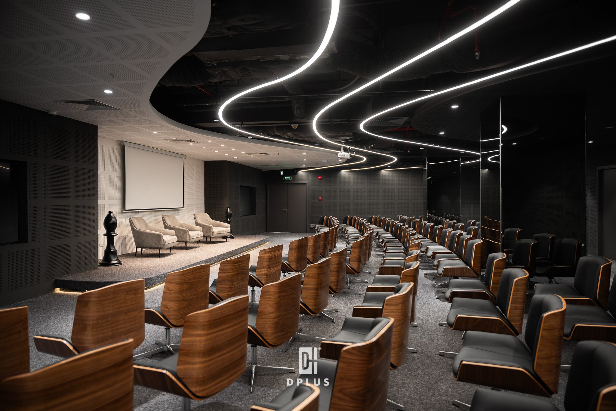 A conference or seminar room with a modern design, featuring a curved ceiling with LED strip lighting and black wall panels. There are several rows of wooden chairs with black seats arranged facing a stage area with three beige armchairs, side tables