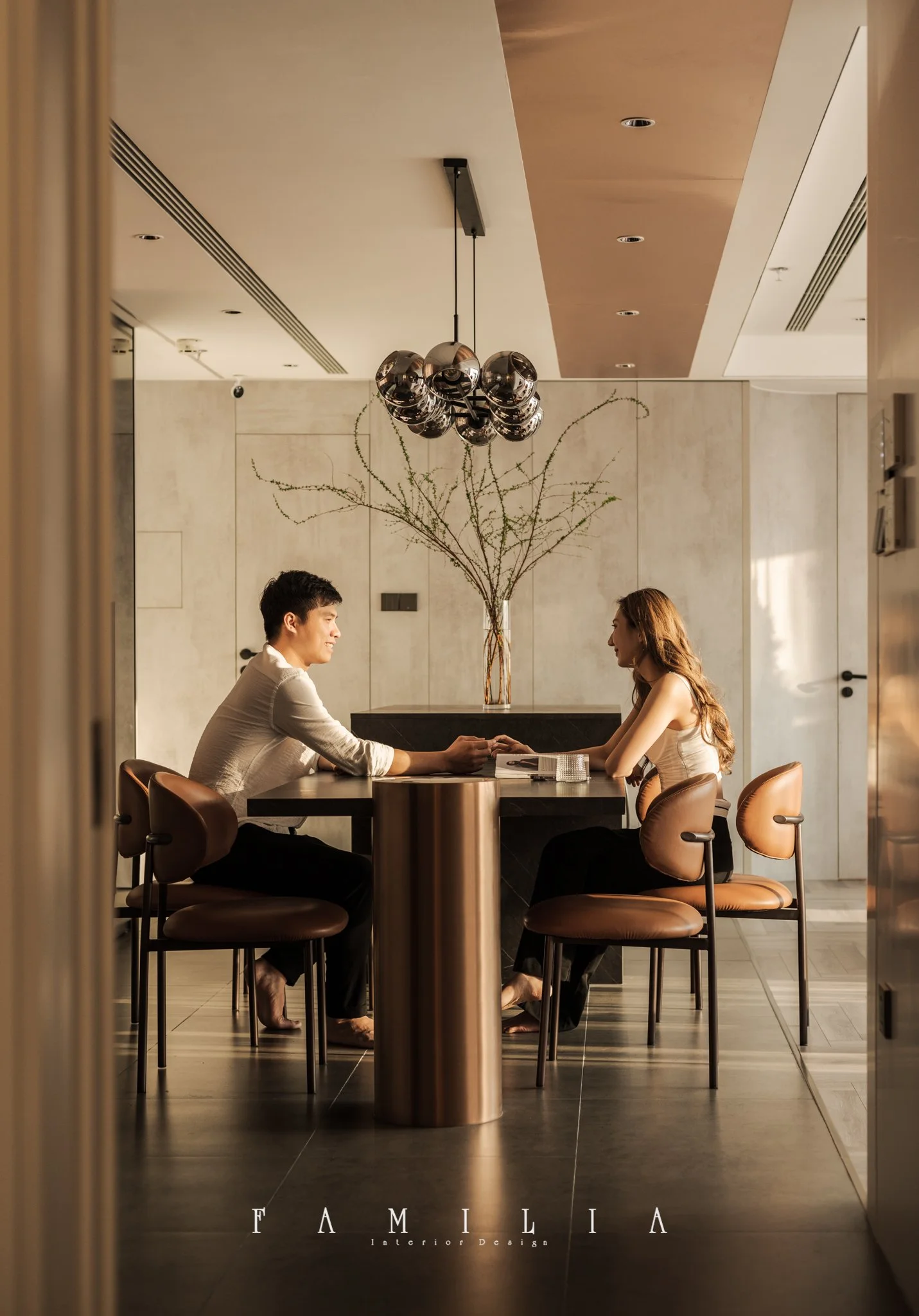 A man and woman sit at a table in a modern, minimalist interior, holding hands and gazing at each other. The room features a decorative vase with dried branches, stylish lighting fixtures, and a sleek, neutral-toned design.