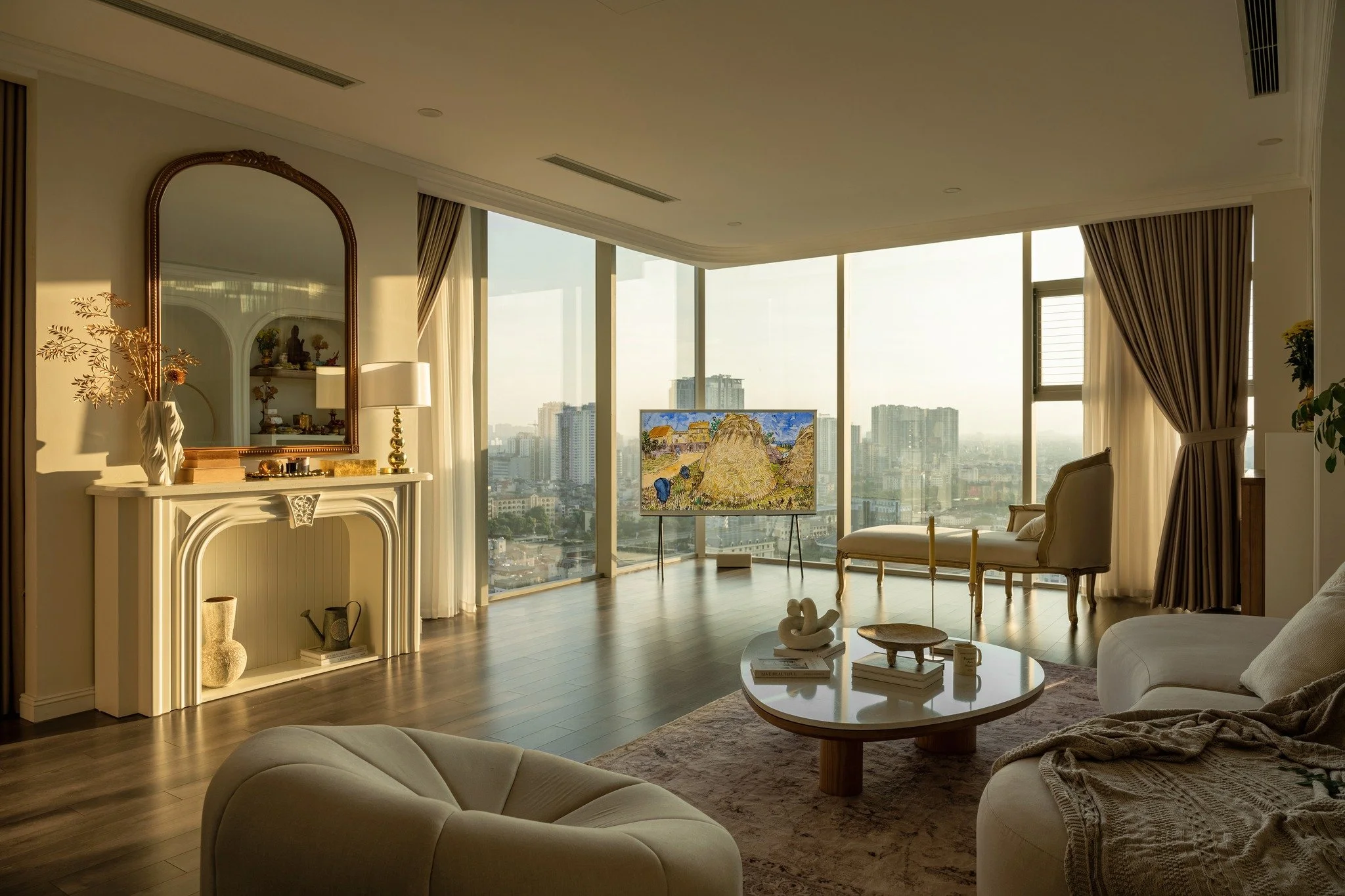 Interior of a modern high-rise apartment living room with large floor-to-ceiling windows showing a city skyline. The room is decorated with elegant furniture, a fireplace mantel with a mirror and decorative items, a round coffee table, and a TV displ