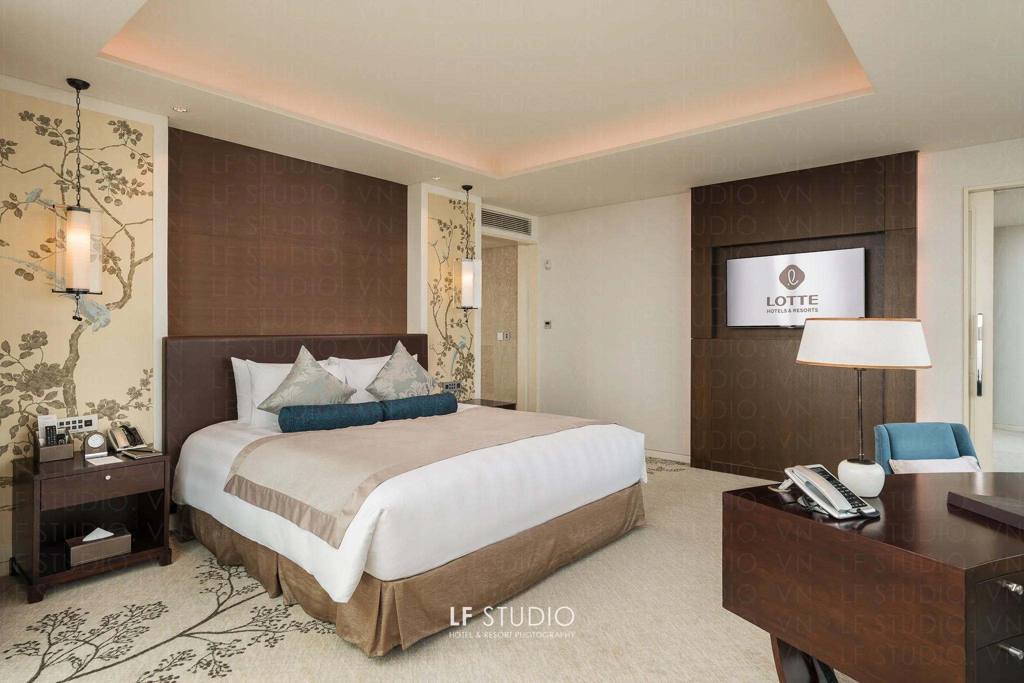 Interior of a hotel room with a bed, bedside table with phones and clock, lamps, a television on the wall displaying 'Lotte Hotels & Resorts', and a comfortable seating area with a lamp and a phone on the table.