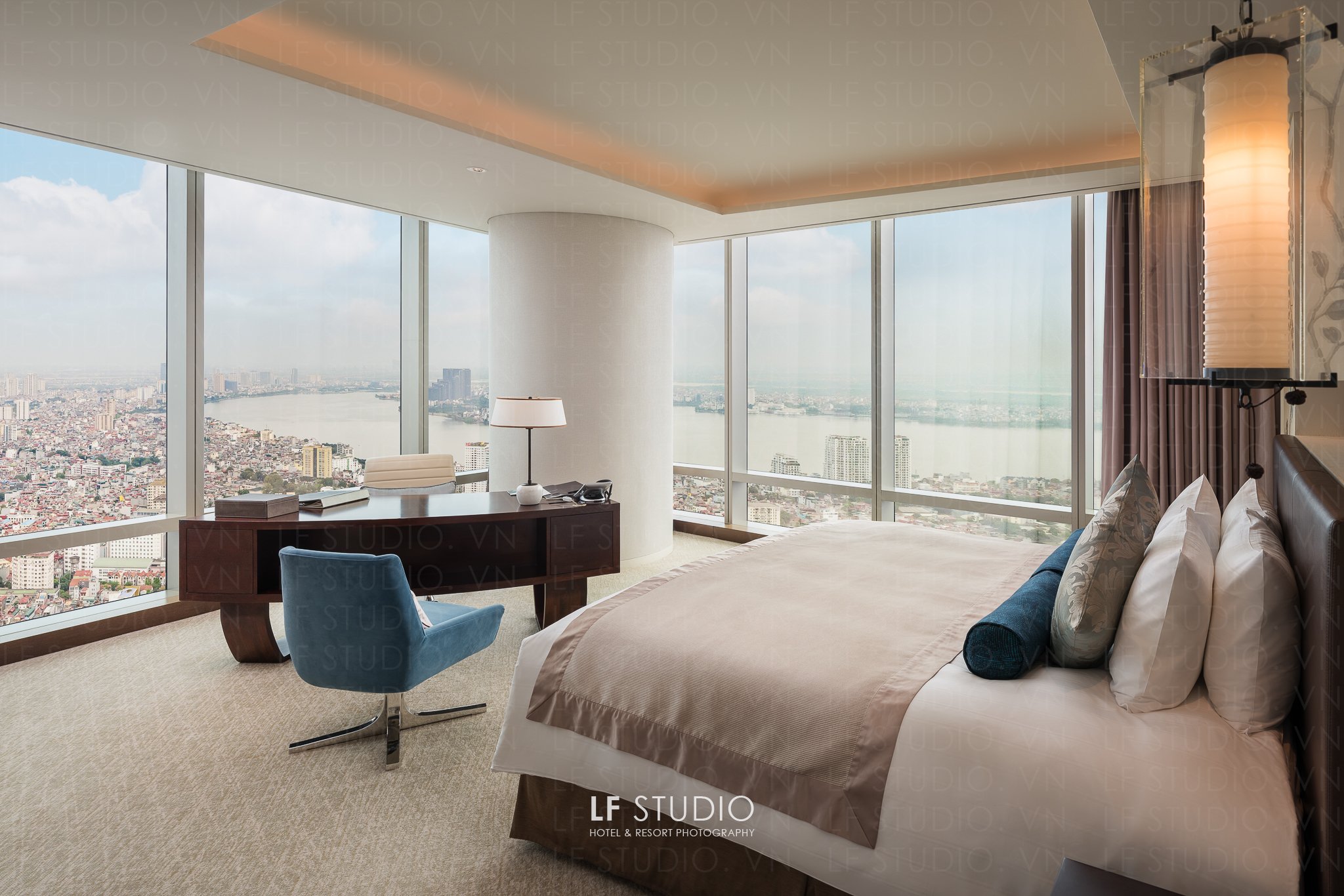 Luxury hotel room with large floor-to-ceiling windows providing a cityscape view of a river and city buildings, a bed with pillows, a bedside lamp, a desk with a phone and books, and a blue chair.