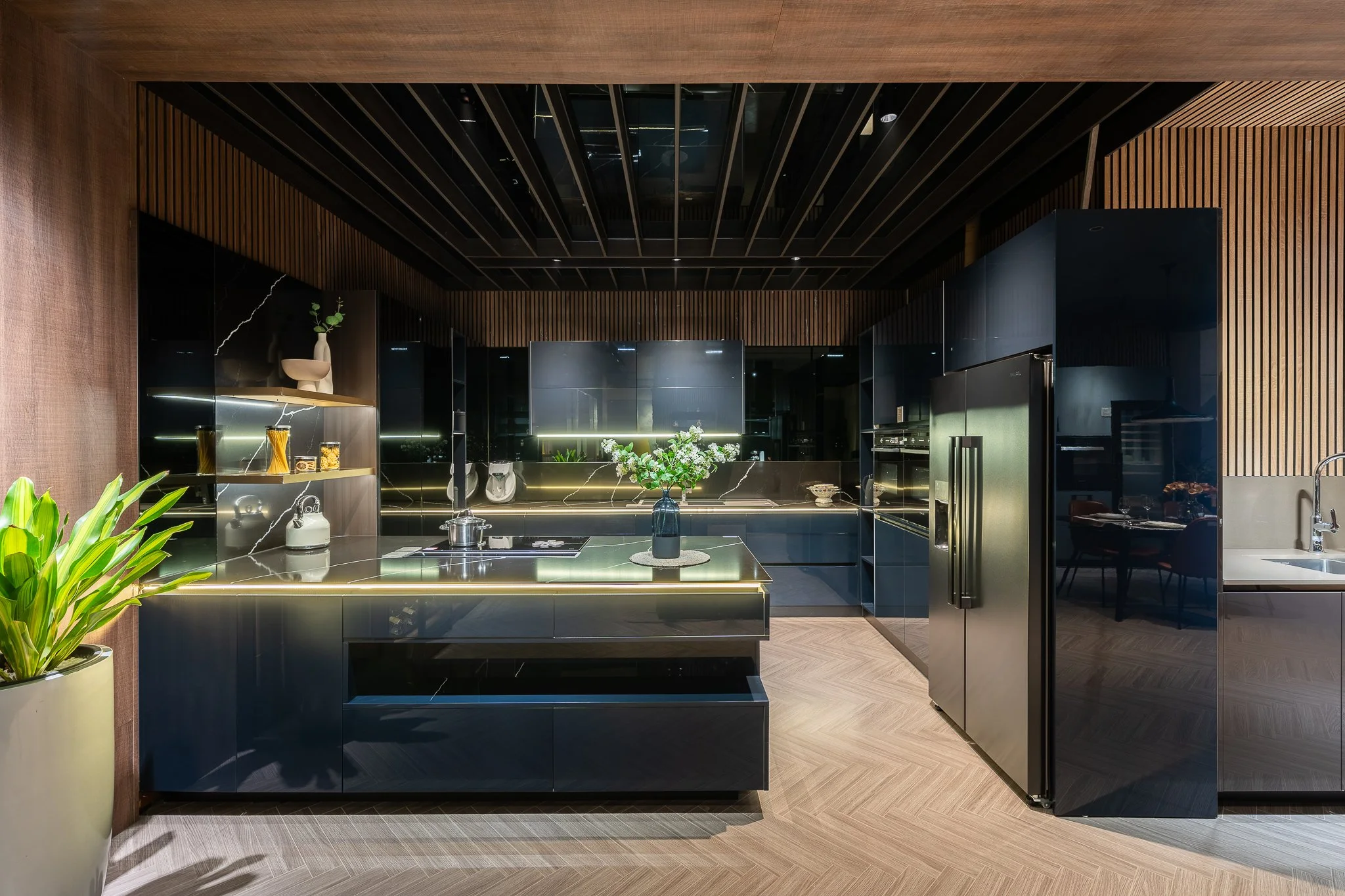 Modern kitchen with black cabinetry, a black marble island with a flower arrangement, wooden ceiling and walls, and a refrigerator on the right side. Plant in a large yellow pot in the foreground.