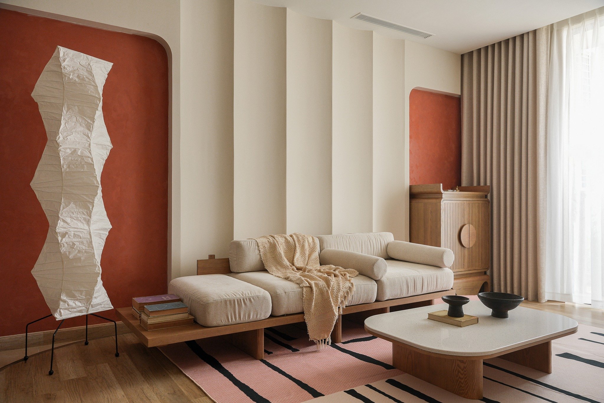 A modern living room with a beige sofa, patterned throw blanket, wooden coffee table with black and beige bowls, tall white lamp, and built-in wooden cabinet near a large window with beige curtains. The wall behind the sofa is painted terracotta.