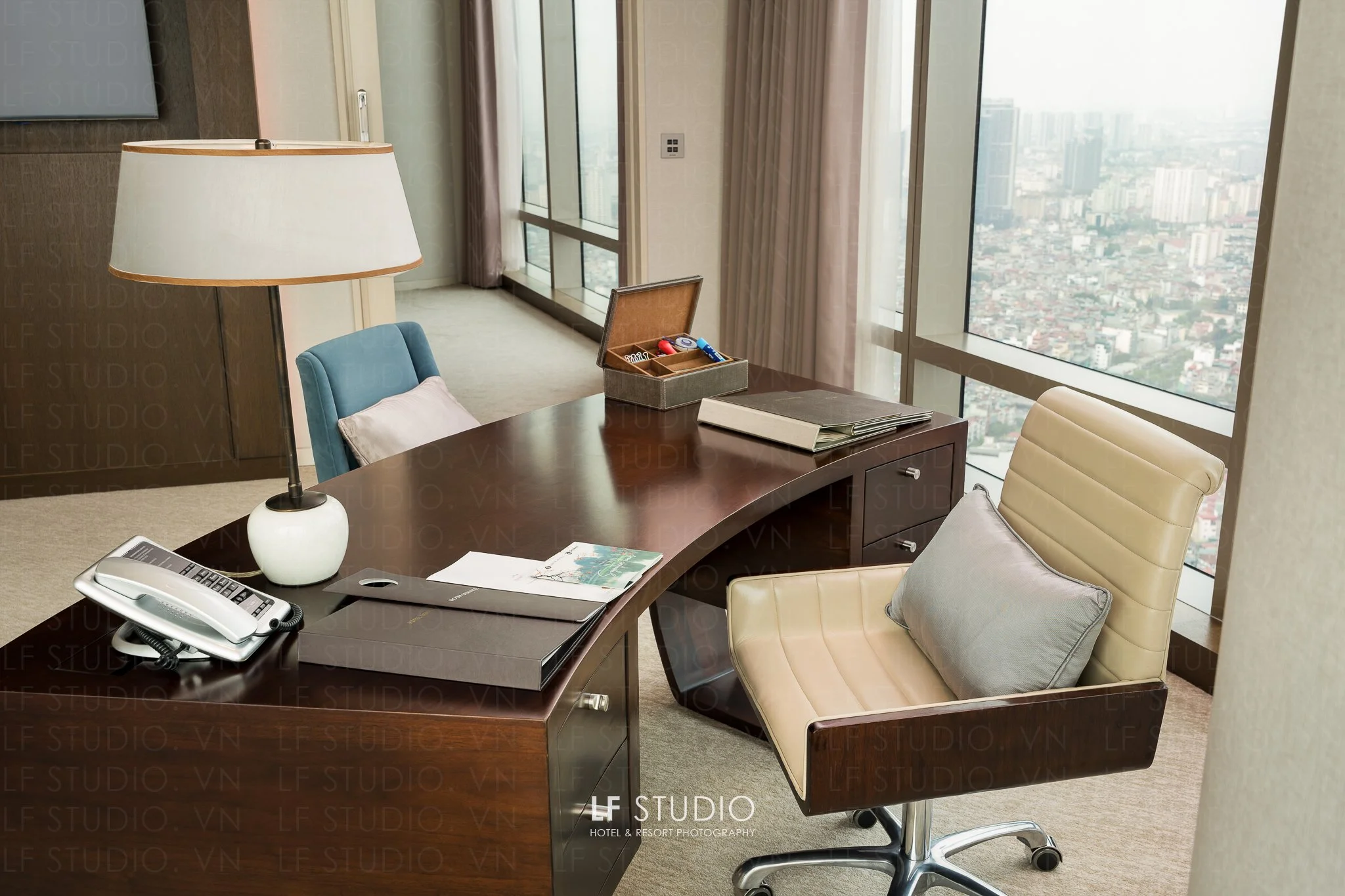 A hotel conference room or office with a large wooden desk, beige and teal chairs, a white desk lamp, a telephone, a book, and a box with small items on the desk. There are floor-to-ceiling windows with cityscape views, curtains, and a control panel on the wall.