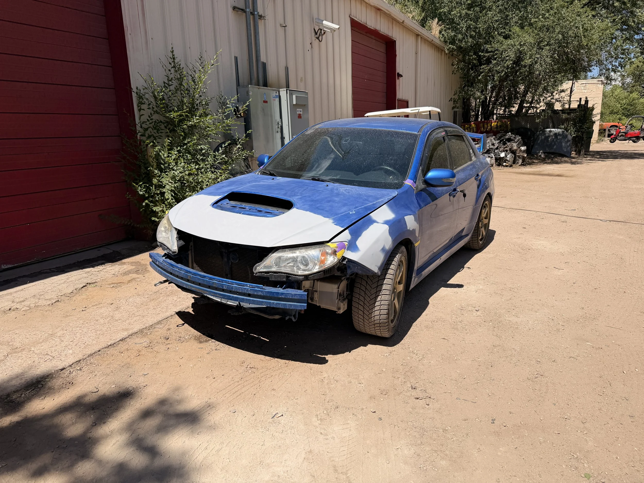 A blue car with a damaged front bumper and hood parked outside a red and beige building, with dirt ground and trees in the background.