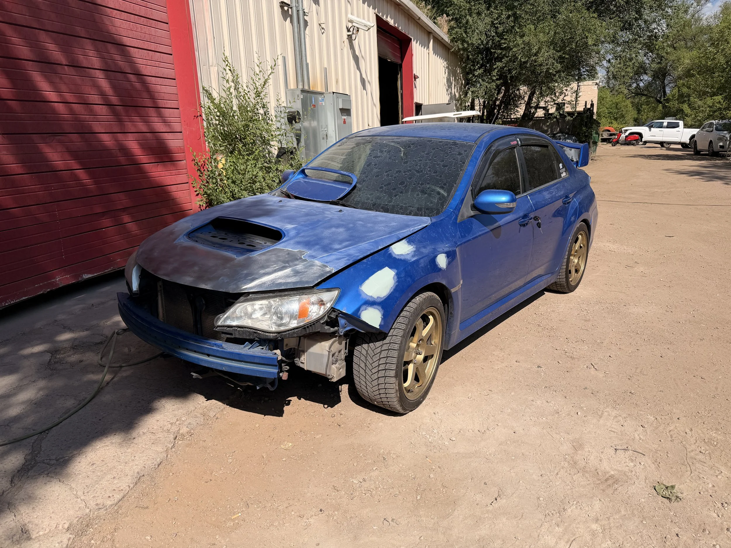 A blue car with a missing front bumper, revealing mechanical parts below, parked on a dirt lot outside a building with beige and red walls. The car has signs of wear and some dents, with the hood and windshield dusty and some spots of paint damage. In the background, there are several other vehicles, trees, and utility boxes.