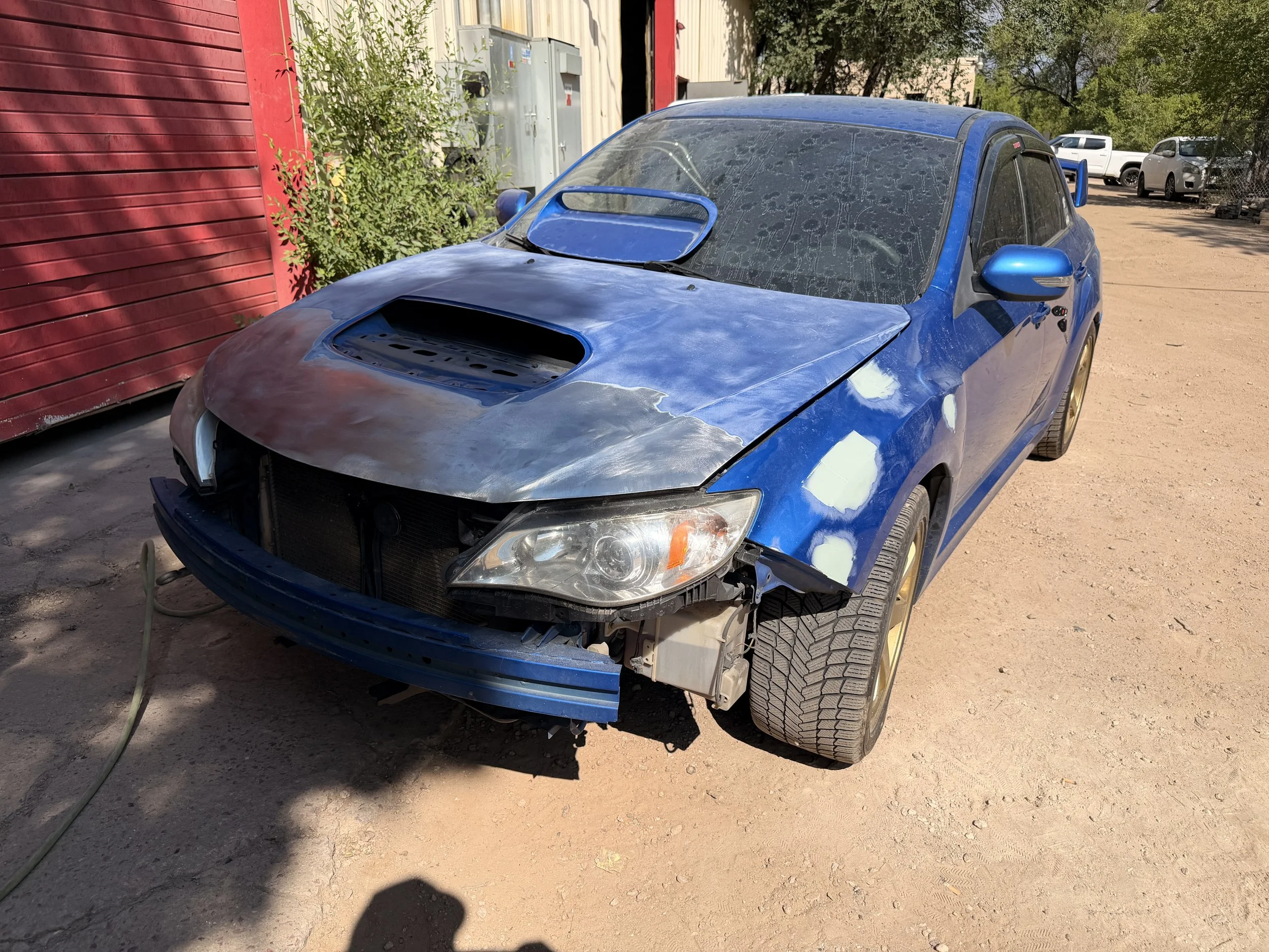 Blue car with front bumper partially removed and hood raised, parked on dirt next to a red building and green bushes, with other vehicles in the background.