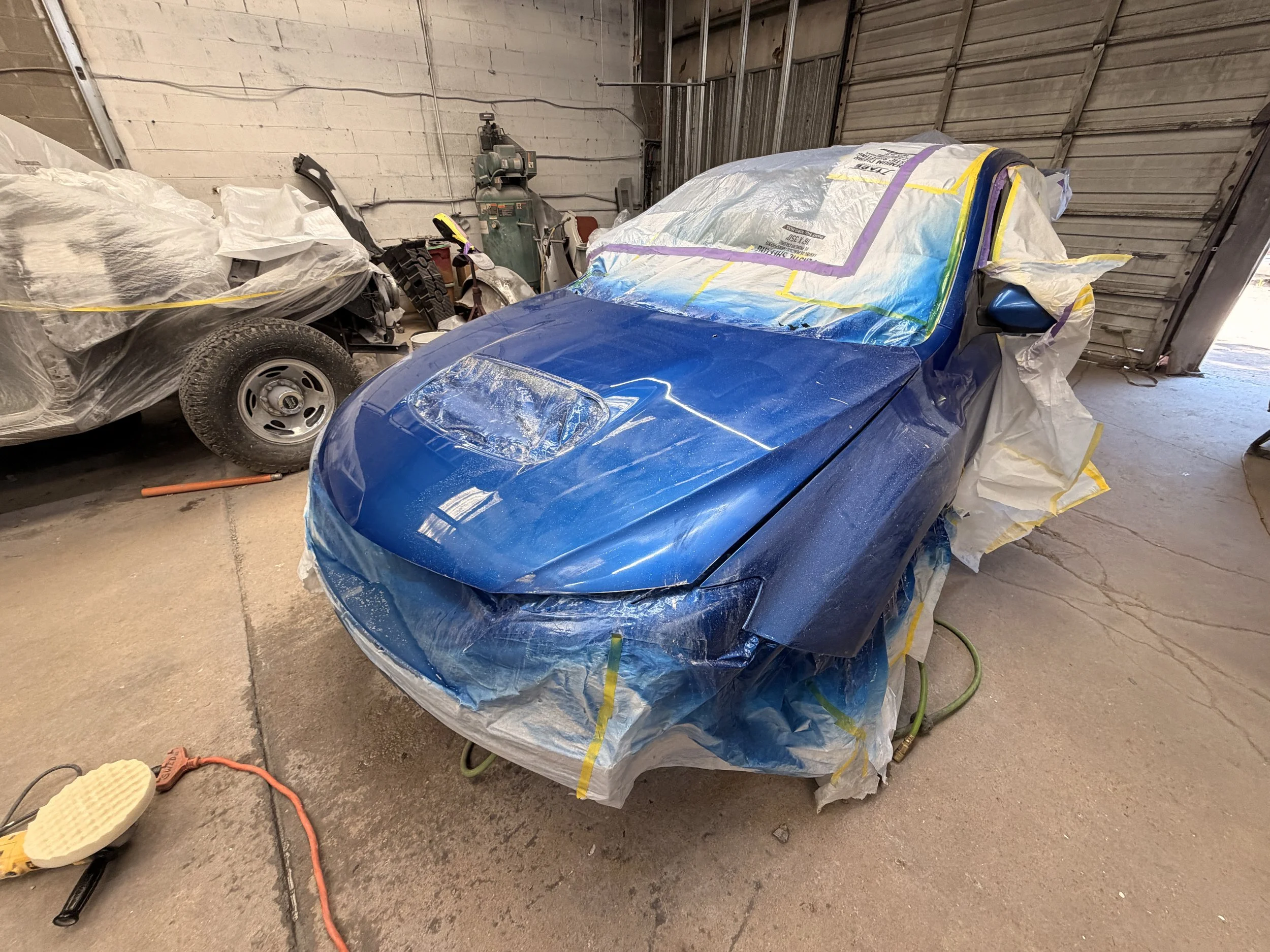 Unmarked blue sports car covered with painter's paper and tape in a garage or workshop. Nearby is a car with damage, rust, and a flat tire, along with various tools and equipment.