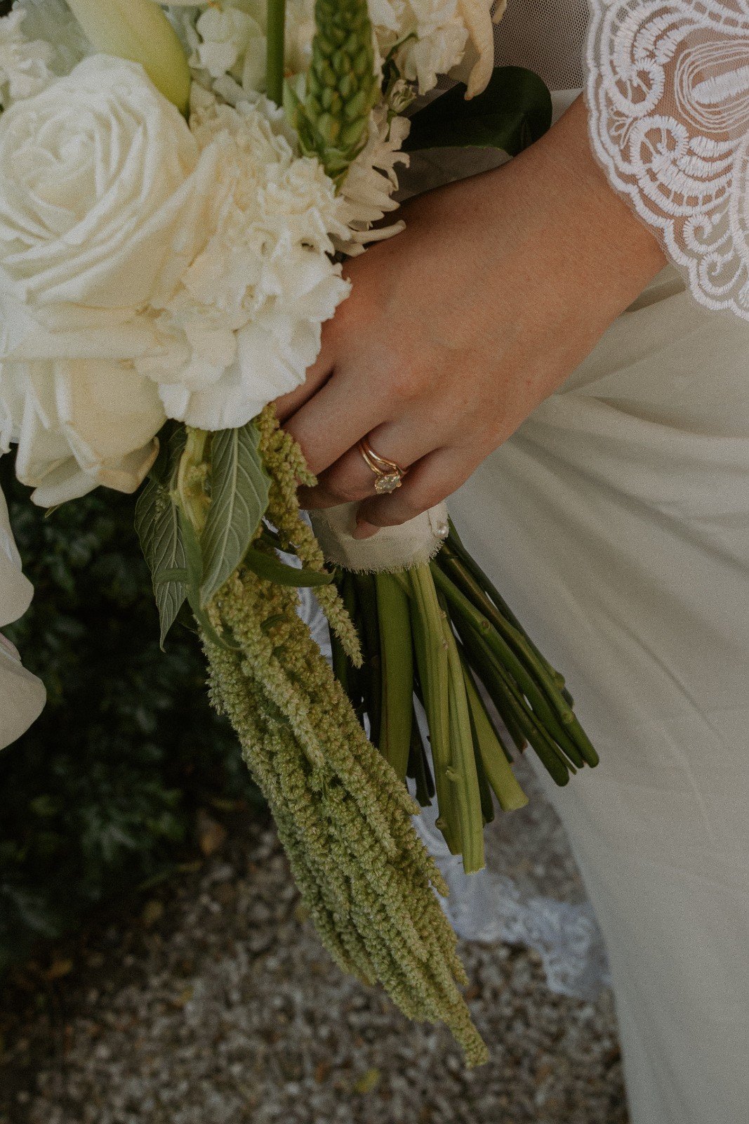 Close-up of a woman's hand with an engagement ring, holding a bouquet of white flowers including roses, hydrangeas, and greenery, with lace detail on her sleeve.