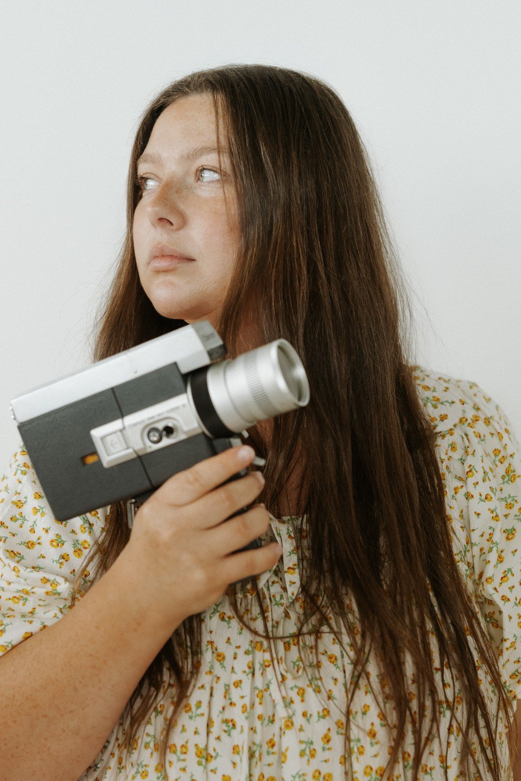 Robyn Small with long brown hair holding a vintage camera, wearing a floral dress, looking to the side against a plain white background.