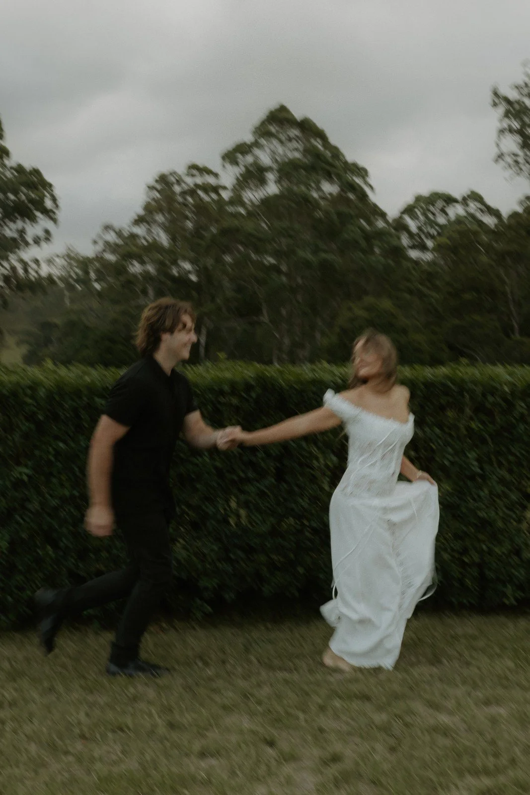 A man in a black shirt and pants holding hands with a woman in a white dress, running outdoors on a cloudy day with trees and bushes in the background.