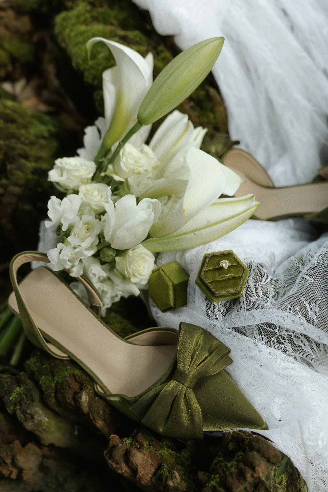 A bouquet of white lilies and small white flowers resting on a lace cloth, with green jewelry boxes, a green bow, and a beige high heel shoe nearby.