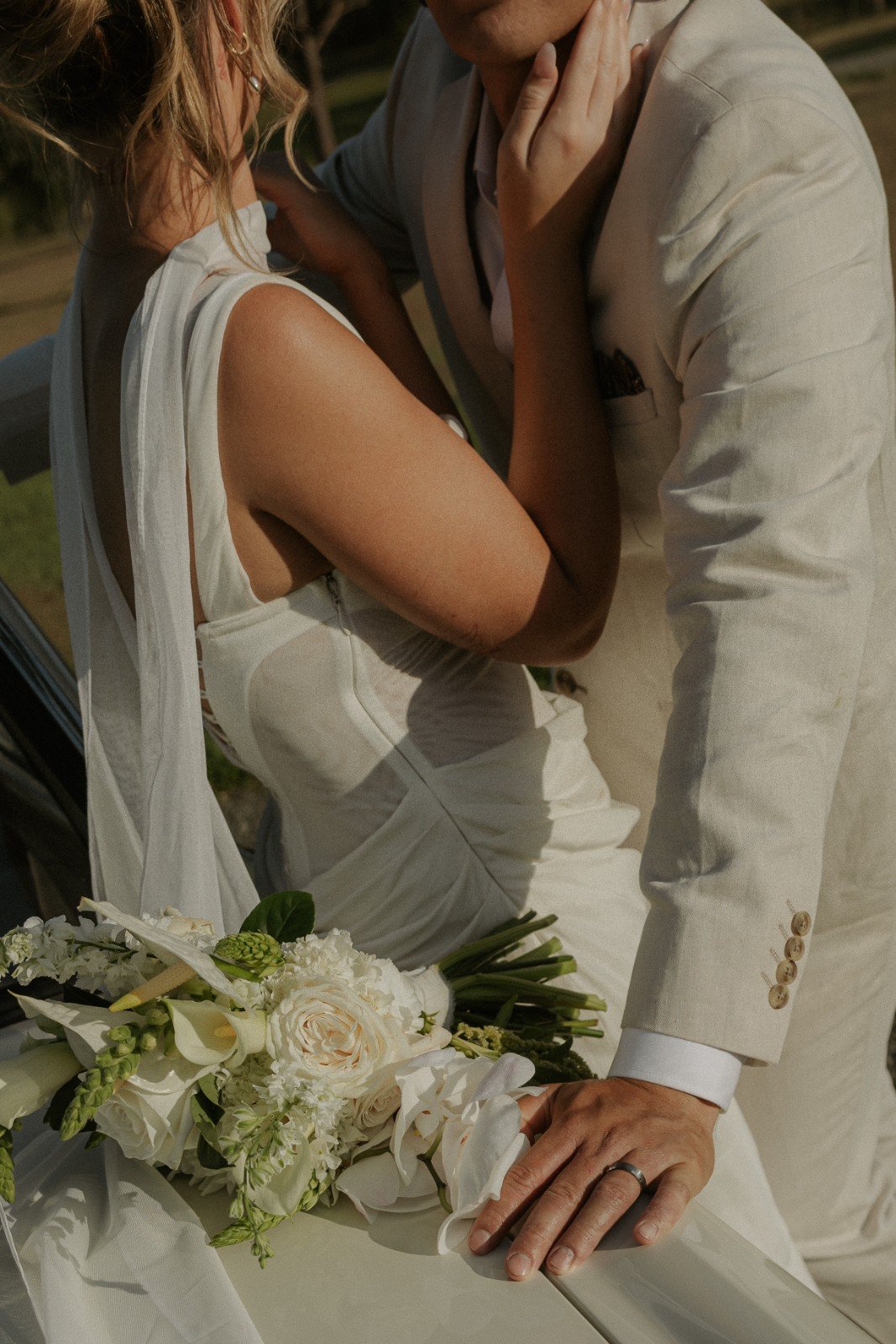 Close-up of a bride and groom in wedding attire, touching foreheads with hands near a bouquet of white flowers, outdoors in daylight.