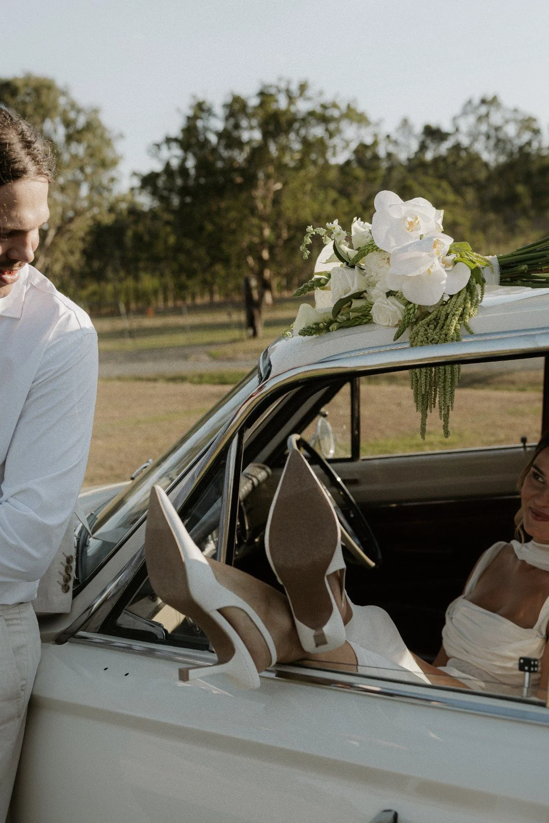 A woman dressed in white leather shoes and a white dress sitting in a vintage white car with a bouquet of white flowers on the roof, outside on a sunny day.