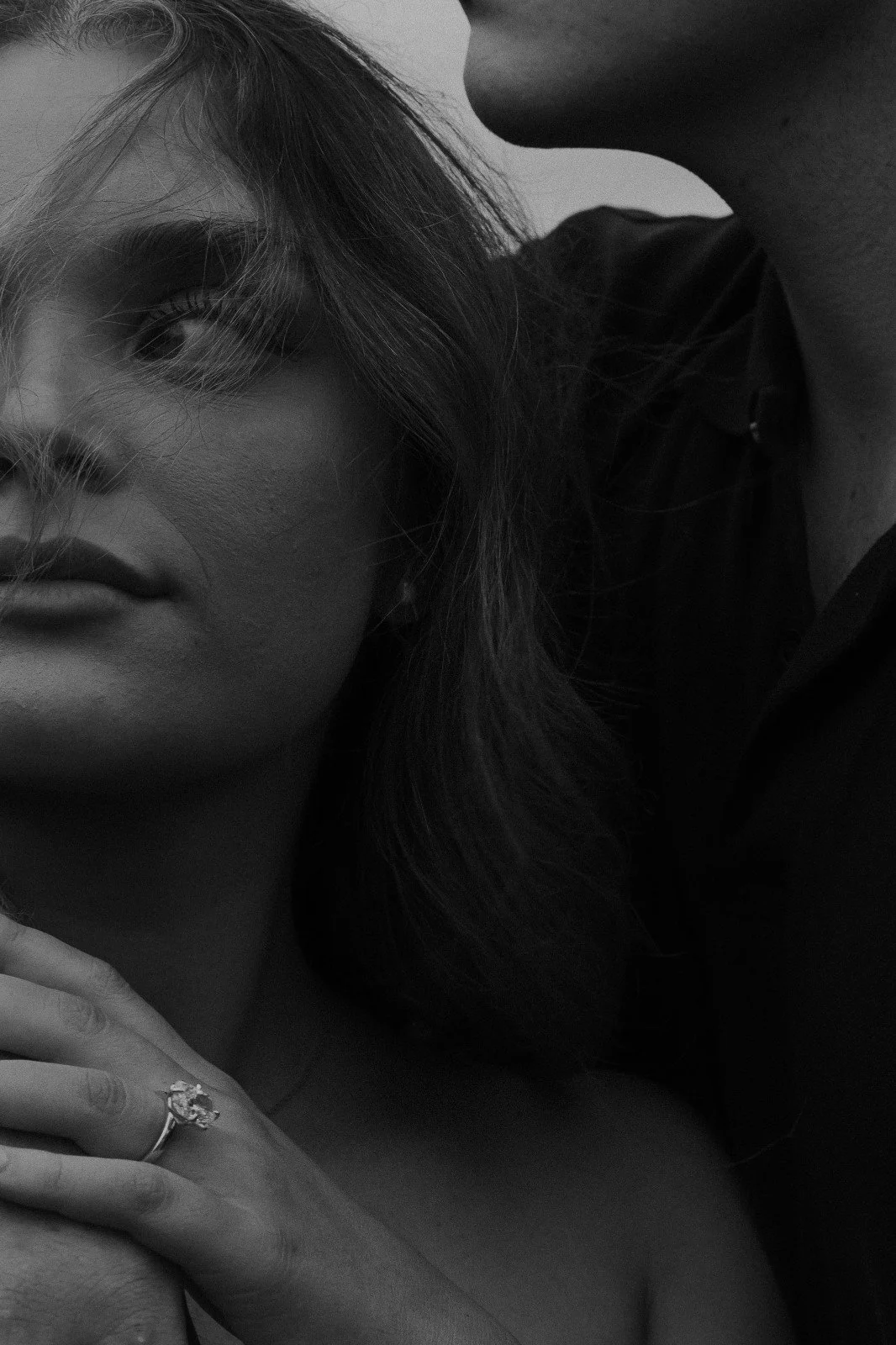 Close-up of a woman with long, wavy hair and eyebrows, holding her hand to her chest, wearing a ring with a gemstone.