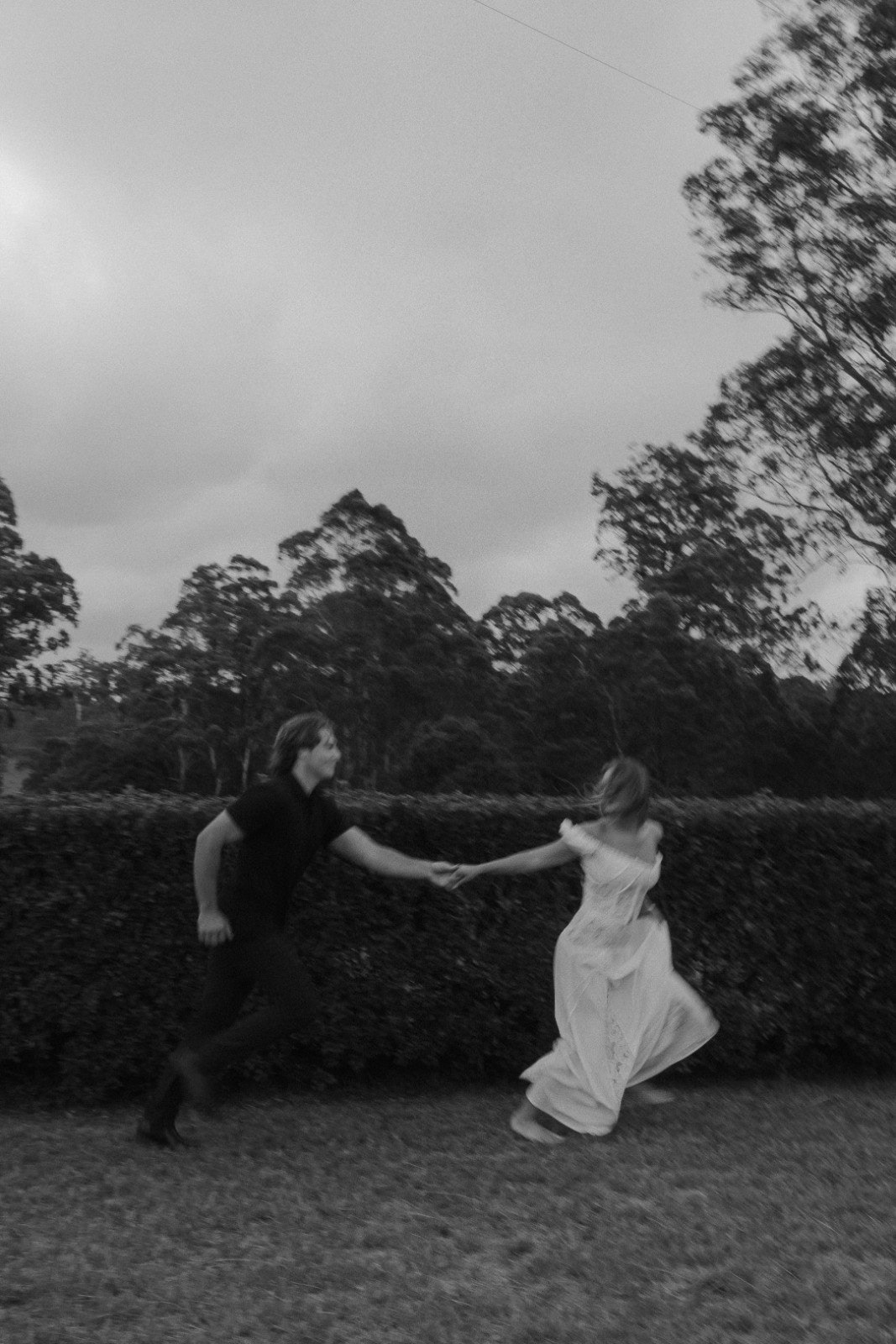 A black and white photograph of a man and woman on their holding hands while dancing outdoors. The man is wearing a dark shirt and pants, and the woman is in a long, flowing dress. They are surrounded by trees and bushes, with a cloudy sky overhead.