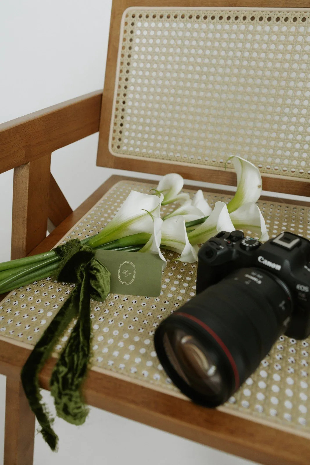 A bouquet of white calla lilies tied with a green ribbon, a black Canon camera, and a wooden chair with a woven cane seat and backrest.