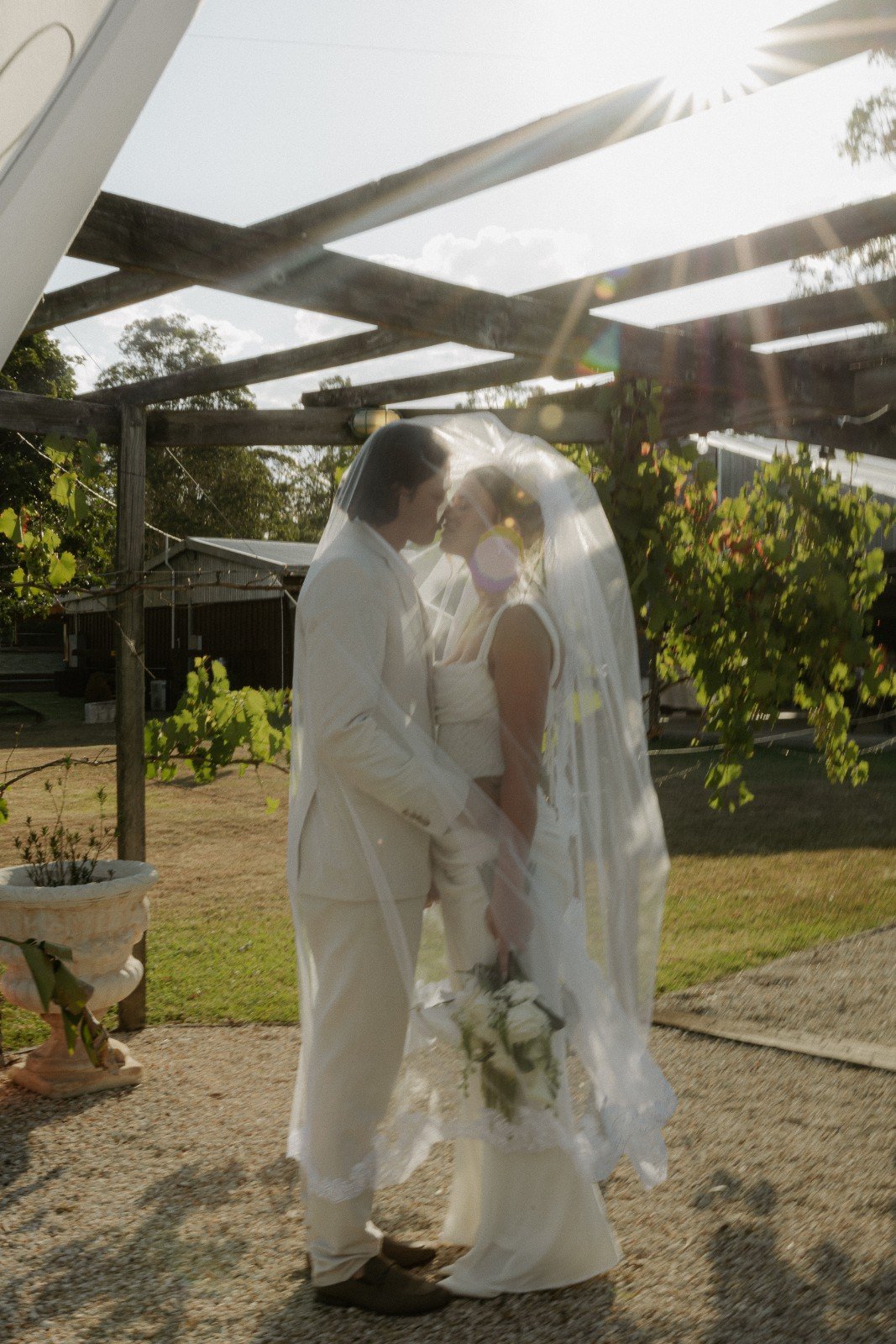 A bride and groom are standing close together under a wedding veil, sharing an intimate moment outdoors in sunlight.