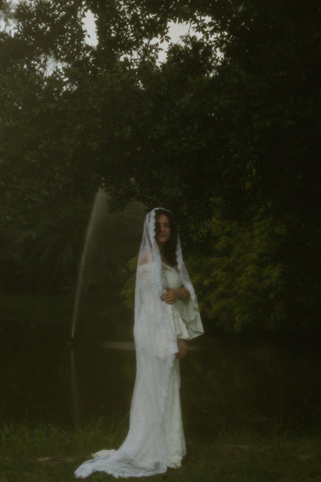 A woman dressed in a white gown and veil standing outdoors under a tree near a water fountain.