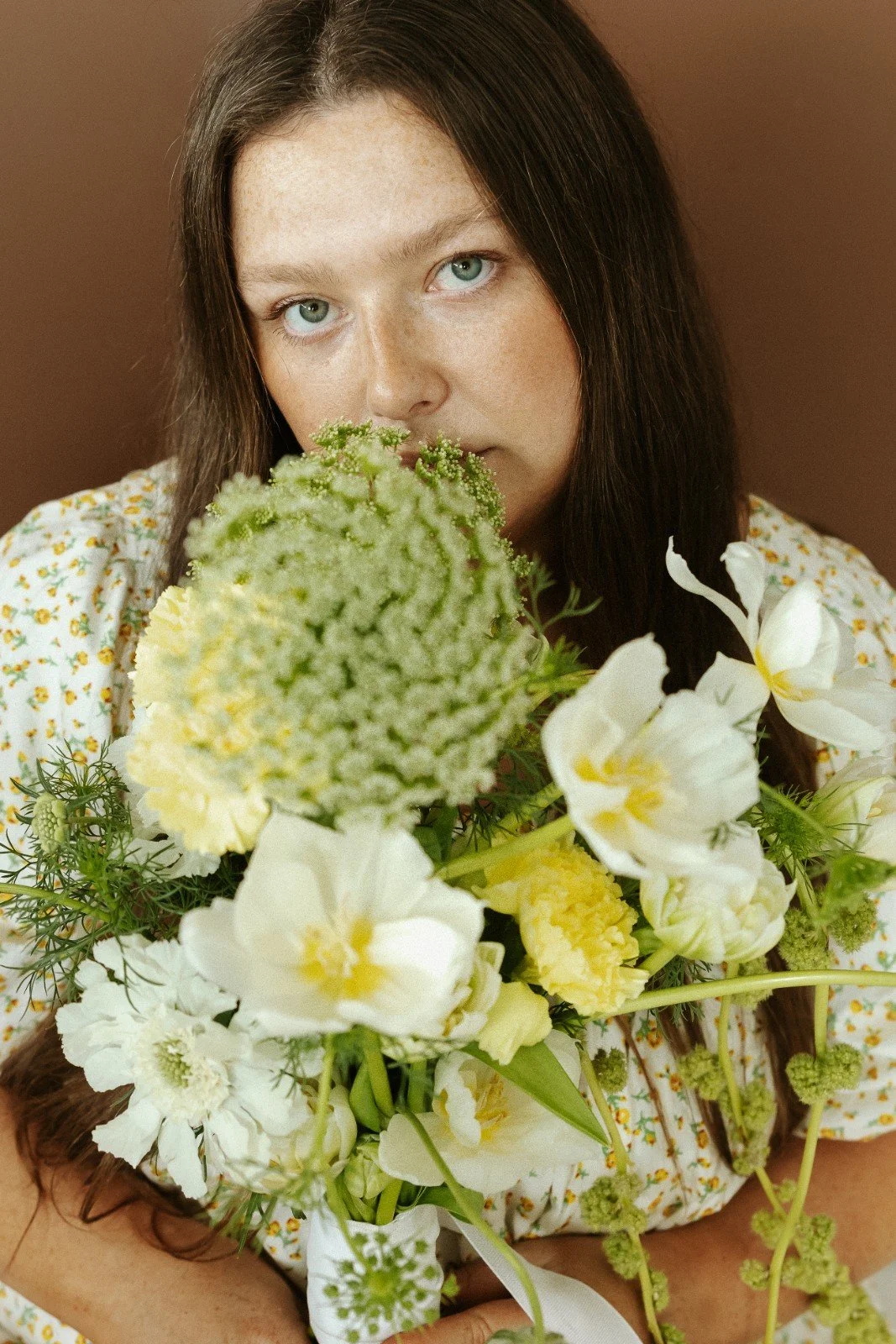 Robyn Small with long dark hair and blue eyes holding a bouquet of white and green flowers against a brown background. Branding for Bloom Film