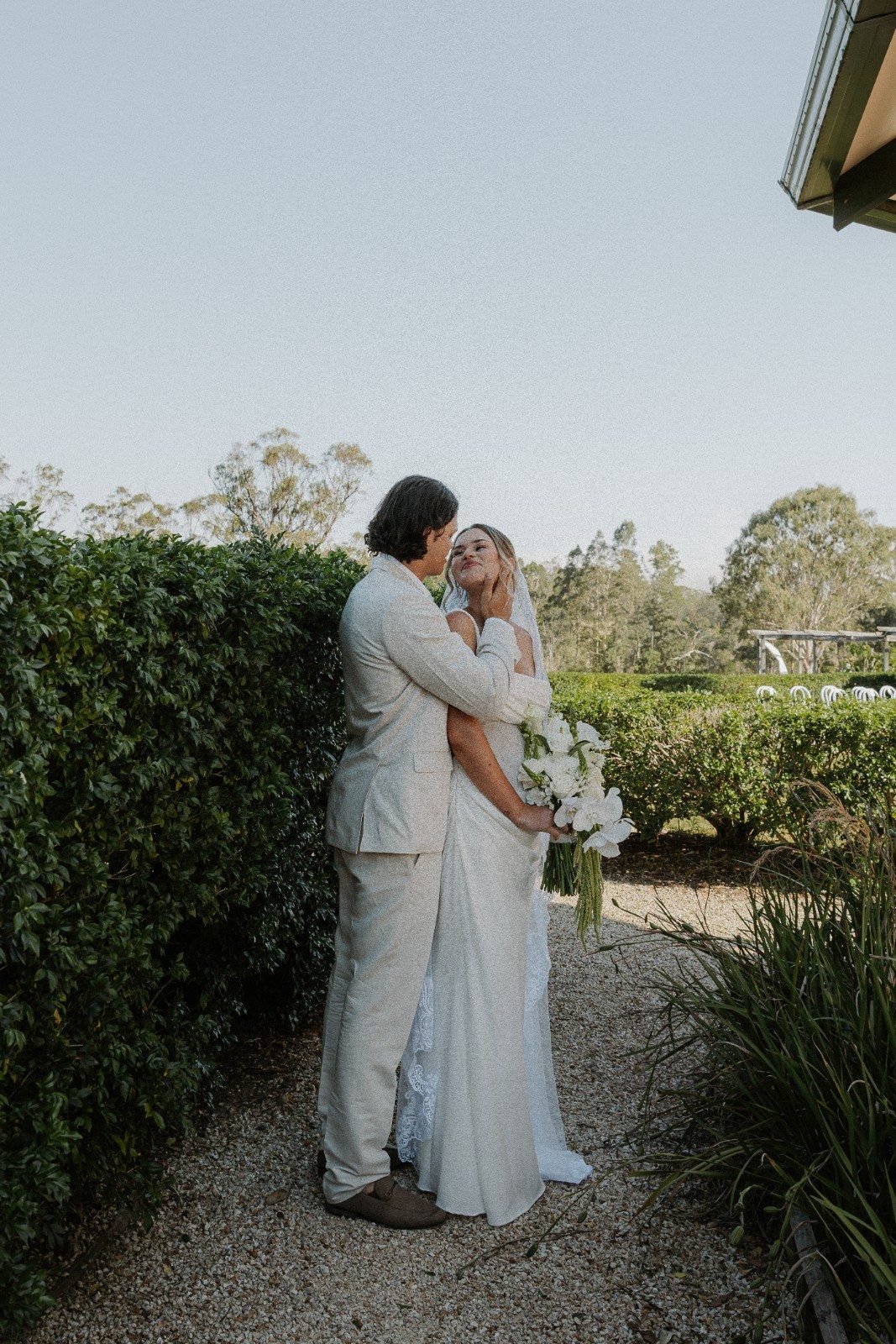 A bride and groom embrace outdoors during their wedding, surrounded by greenery, with the bride holding a bouquet of white flowers.