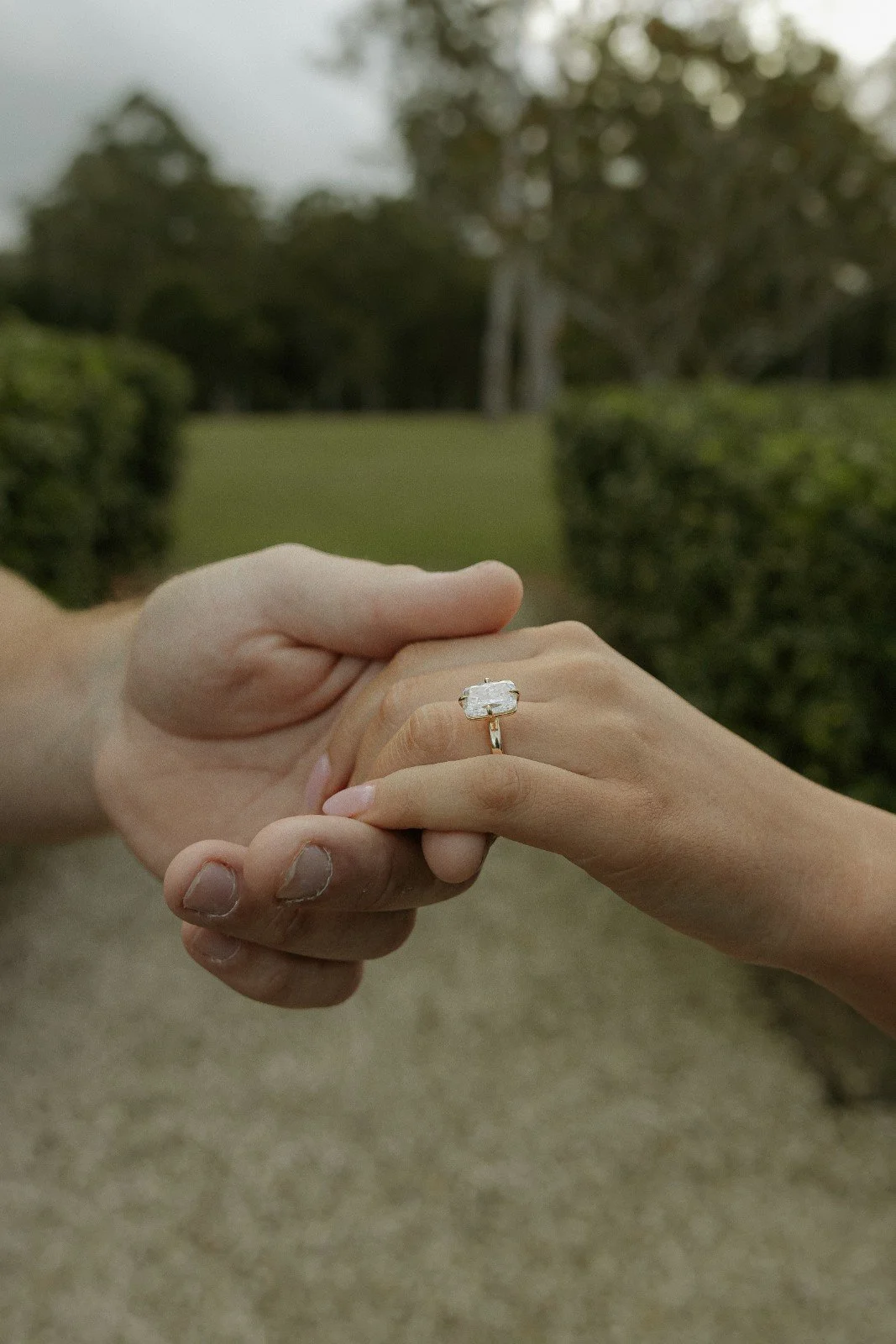 A person's hand with a wedding band holding an engagement ring with a large rectangular diamond in a garden setting.