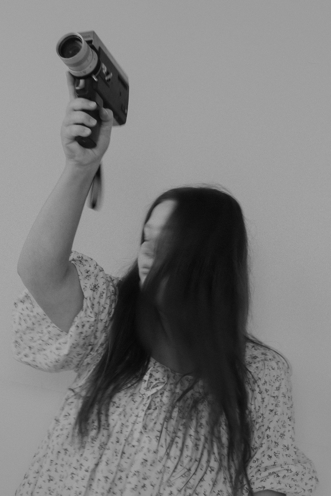 Robyn Small with long dark hair, wearing a patterned dress, holding a vintage camera and pointing it upward, with her face partially obscured.