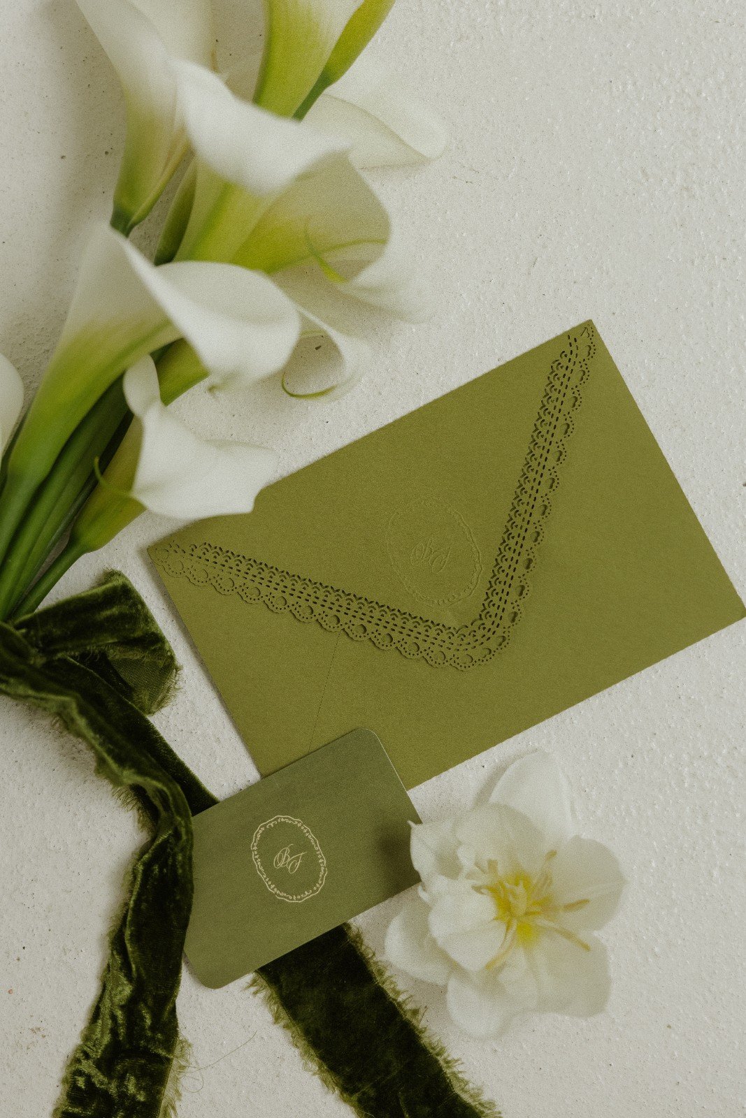 A bouquet of white calla lilies next to a green birthday card with decorative edge and a matching green tag with a monogram, on a white surface.
