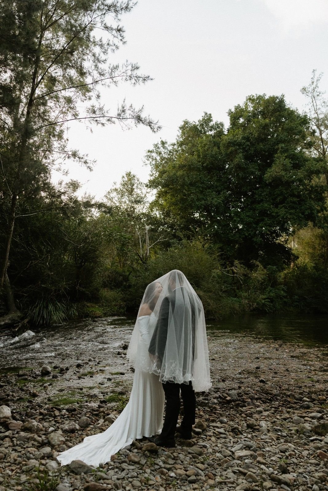 A bride and groom stand together on a rocky riverbank, wrapped in a sheer white veil, with trees and a cloudy sky in the background.