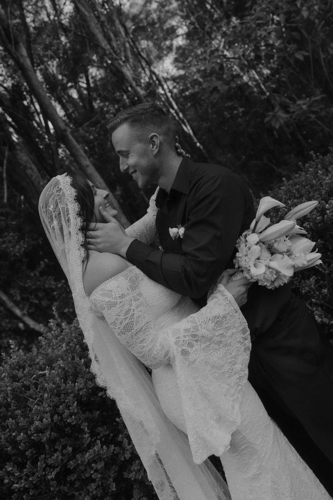A bride and groom sharing a romantic moment outdoors, with the groom holding a bouquet of flowers, both smiling and gazing into each other's eyes.