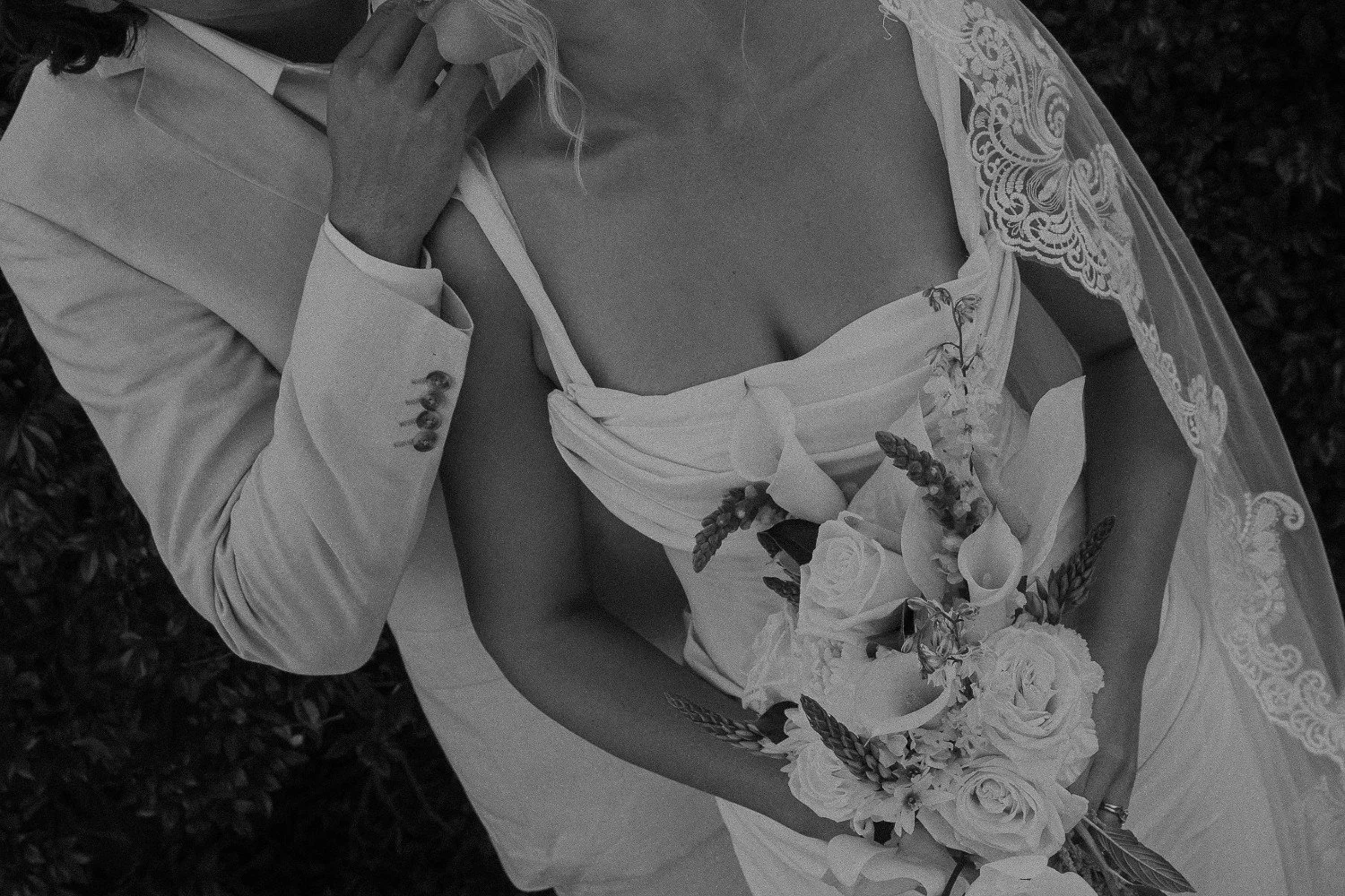 A bride and groom on their wedding day, the bride holding a bouquet of flowers and wearing a wedding gown with lace details on the shoulder, while the groom is in a suit, holding a small object near his face.