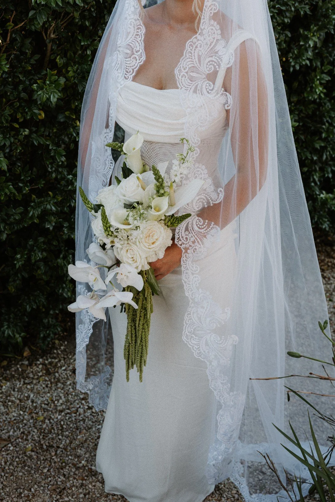 Bride in a white wedding gown holding a bouquet of white roses and calla lilies, standing outdoors in front of dense green hedge, wearing a lace-edged veil.