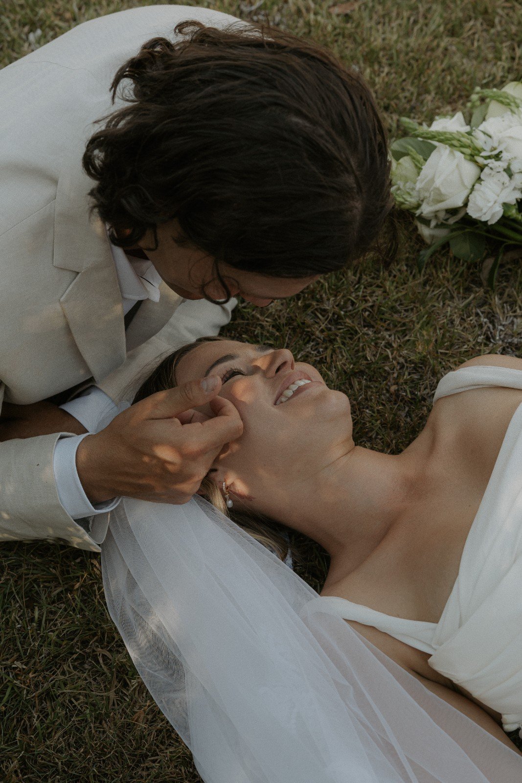 A man in a white suit is kissing a woman lying on the grass in a wedding dress, who is smiling.