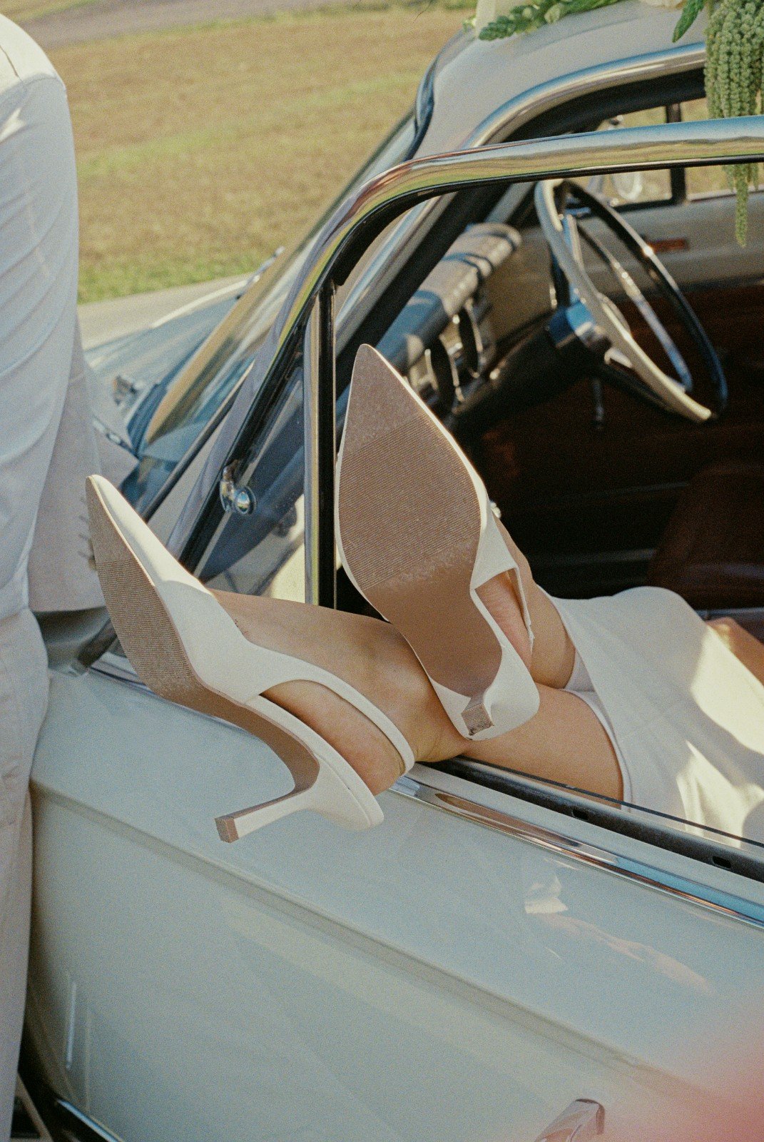 A woman wearing white high heels and a white dress lying with her feet up on the open window of a vintage white car.