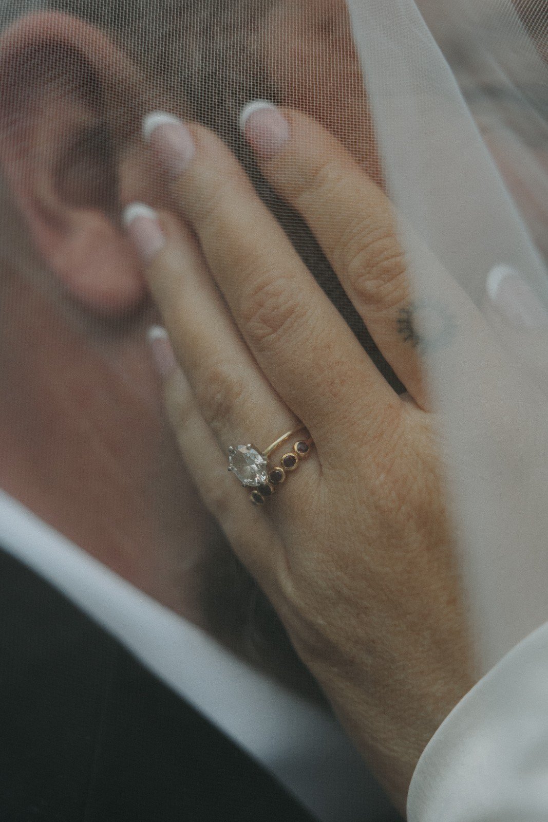Close-up of a hand with a large gemstone ring and small black stone rings, behind a mesh screen.