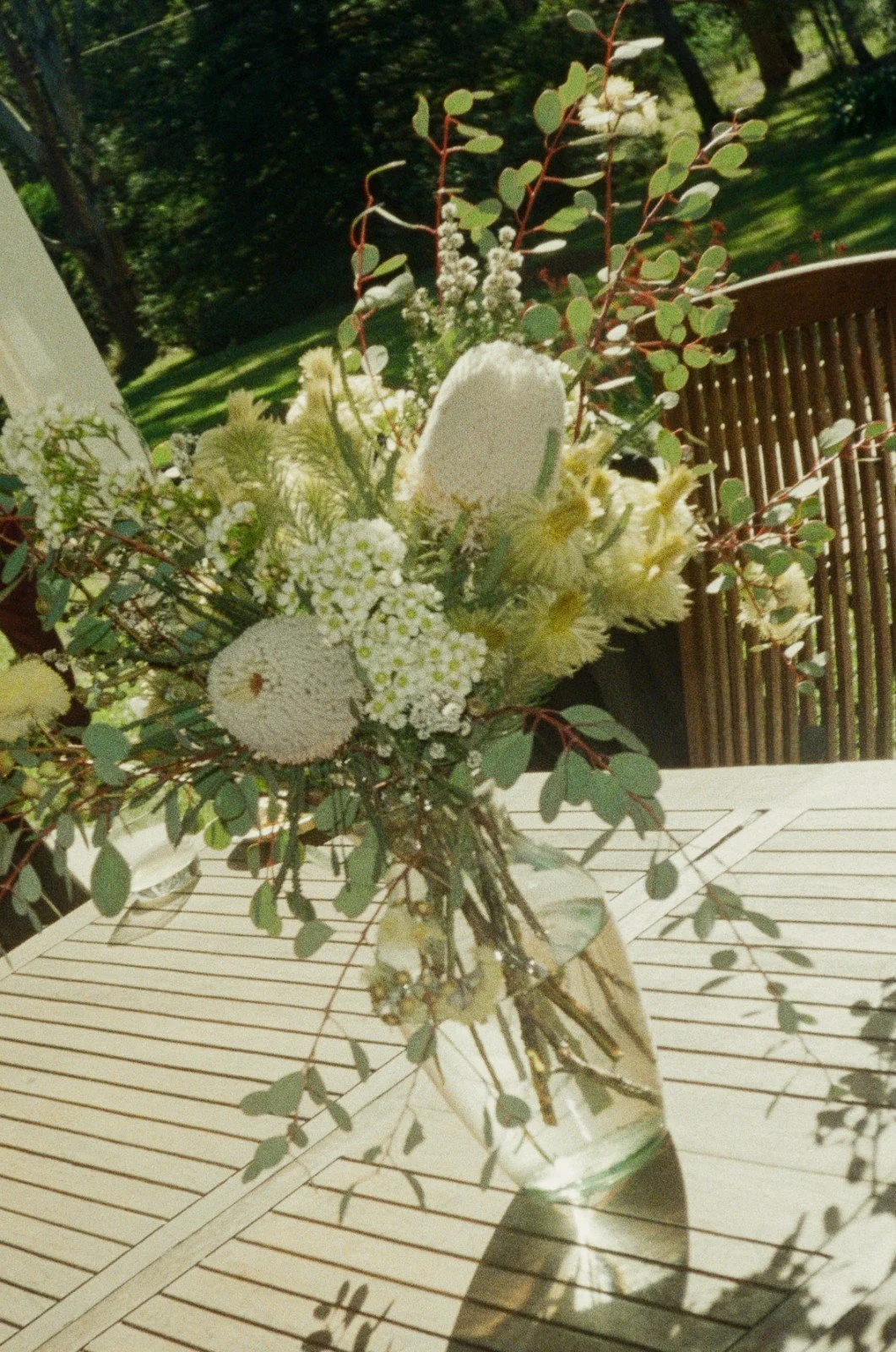 A floral arrangement in a clear glass vase on a white table outdoors, featuring white flowers, eucalyptus leaves, and greenery.