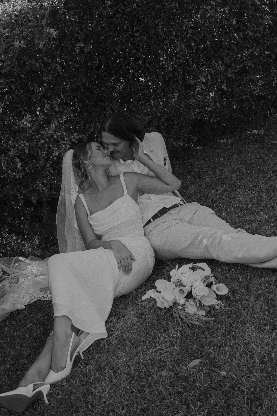 Black and white photo of a couple sitting on the grass, intimate and smiling, with a bouquet of flowers nearby, in front of dense bushes.