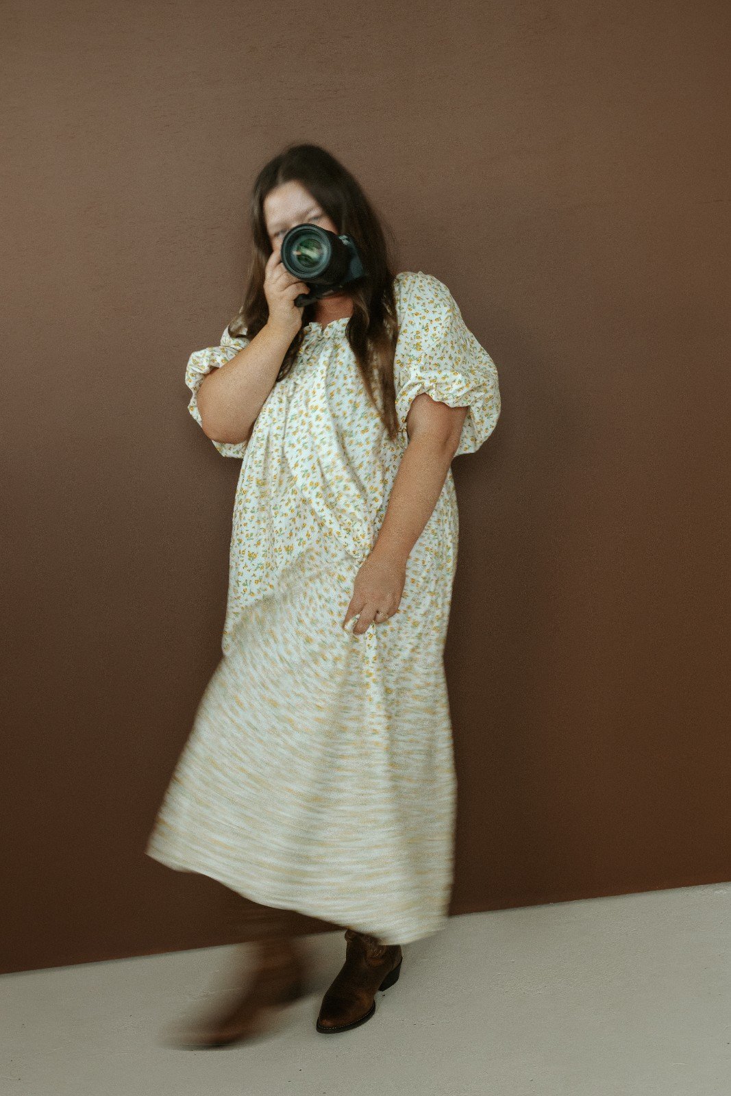 Robyn Small in a floral dress is taking a photo with a camera against a brown wall.